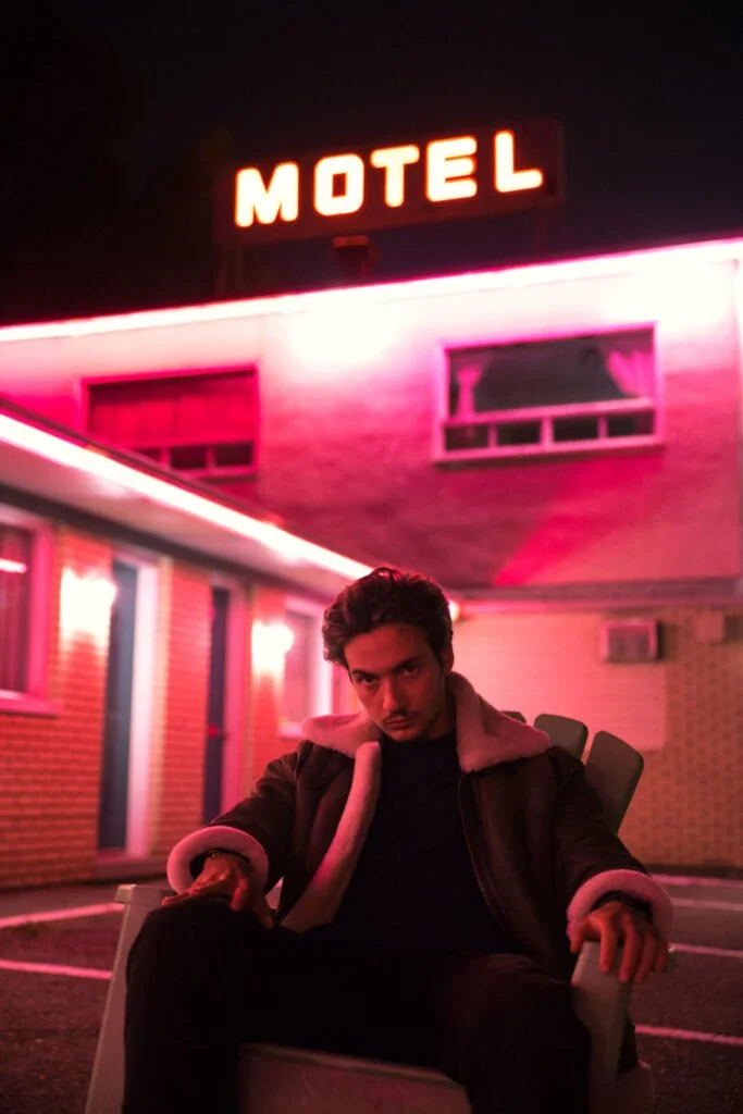 Un homme assis dans une chaise devant un motel avec des néons roses et une enseigne rouge éclairée, la nuit.