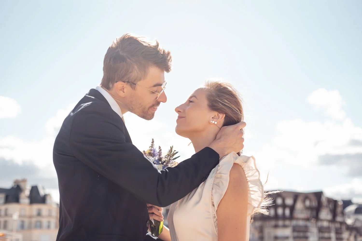 Un homme en costume et une femme sont proches, avec le ciel en arrière-plan, partageant un moment intime.