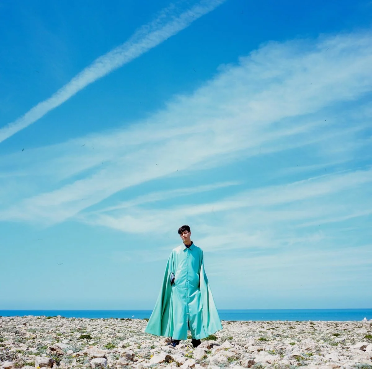 Une personne portant un manteau long turquoise se tient sur une plage rocheuse sous un ciel bleu avec des traînées de nuages. Arthur Avellano
