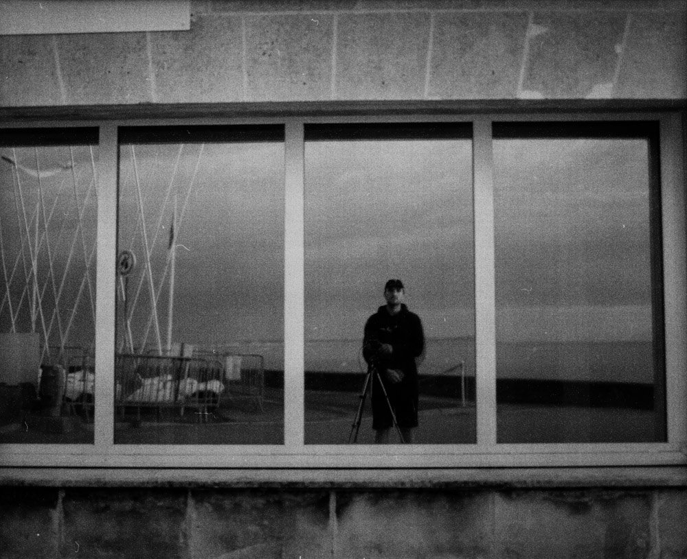 Photo en noir et blanc d'une personne prenant un selfie dans une grande fenêtre, avec un bord de mer visible en arrière-plan.