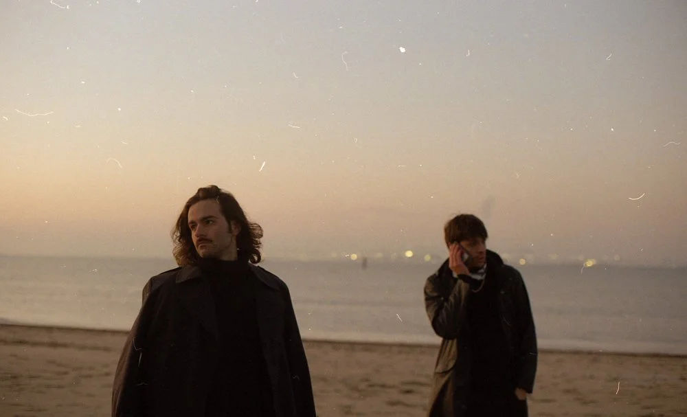 Deux jeunes hommes debout sur une plage au coucher du soleil, one en regardant l'horizon, l'autre à parler au téléphone.