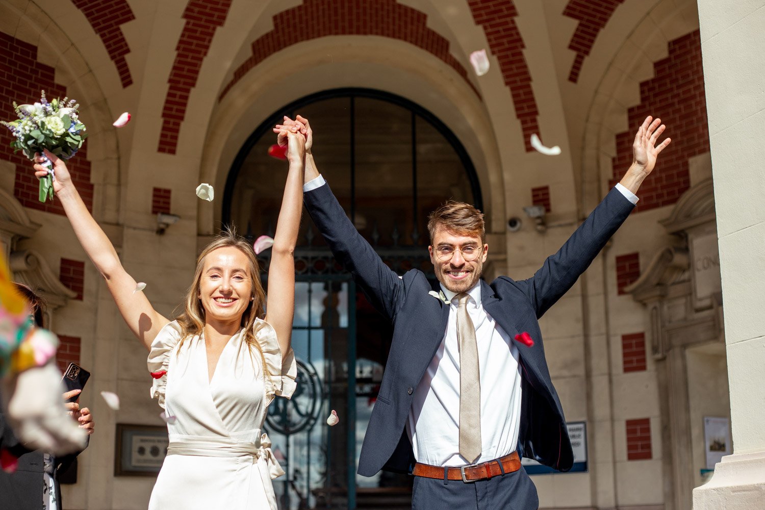 Un couple souriant, homme en costume et femme en robe de mariage, célébrant leur mariage devant un bâtiment archétypal, avec des pétales de rose et un bouquet de fleurs, exprimant leur joie.