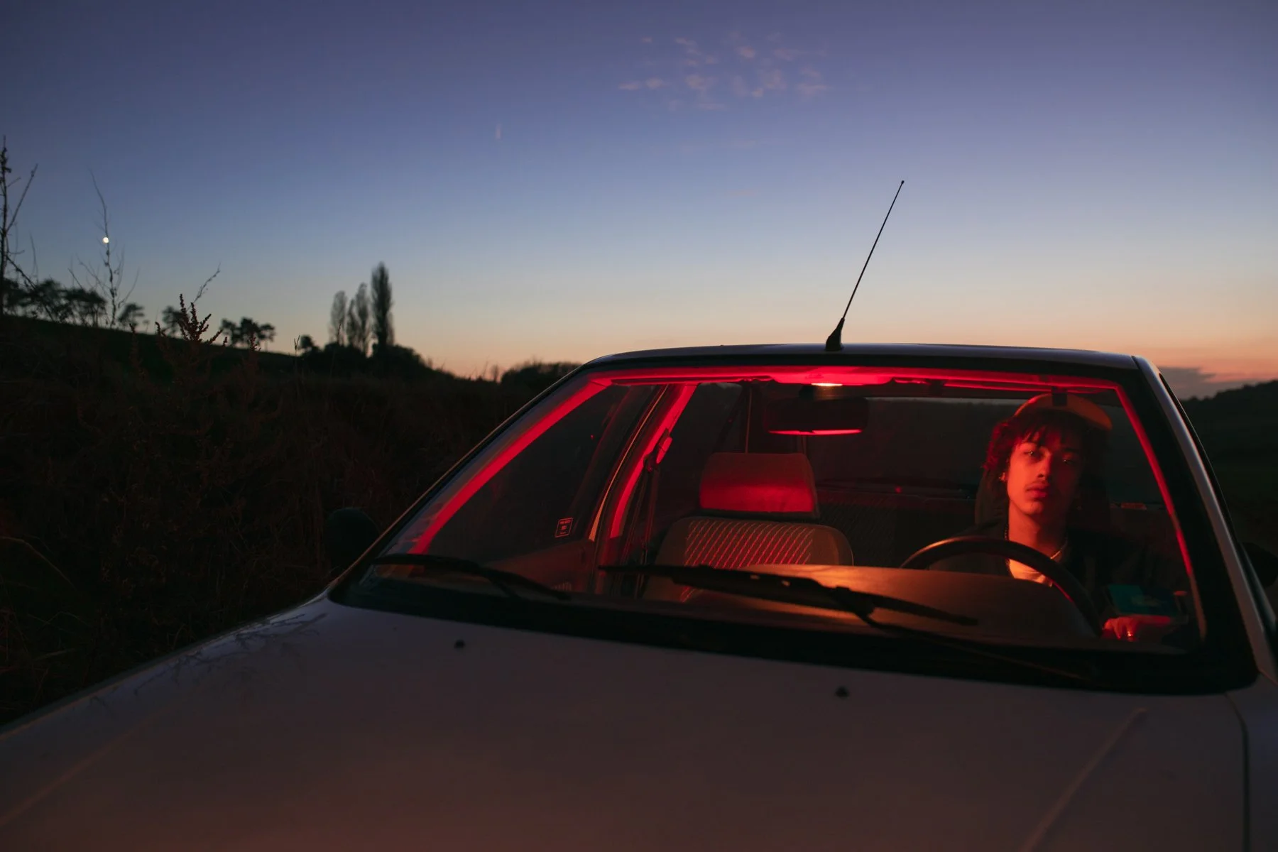 Jeune homme assis dans une voiture au coucher du soleil, avec un ciel coloré et un paysage naturel en arrière-plan.