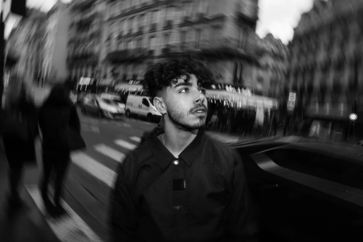 Un jeune homme avec des cheveux bouclés et une barbe, en vêtement sombre, se tient dans une rue urbaine, floue et en noir et blanc, avec des bâtiments, des voitures et deux autres personnes indistinctes en arrière-plan.
