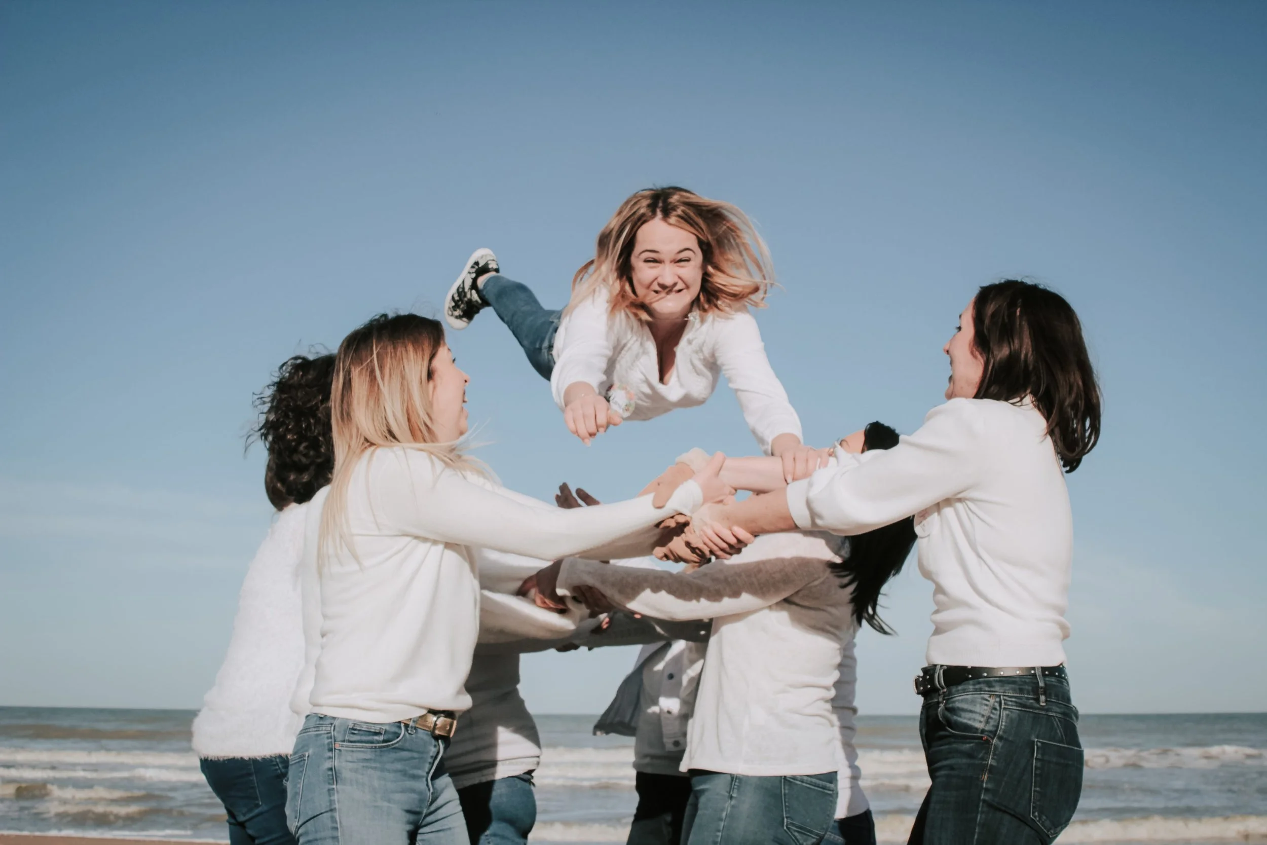 Groupe de personnes en cercle sur la plage, la personne au centre est une femme souriante en train d'être levée par les autres.