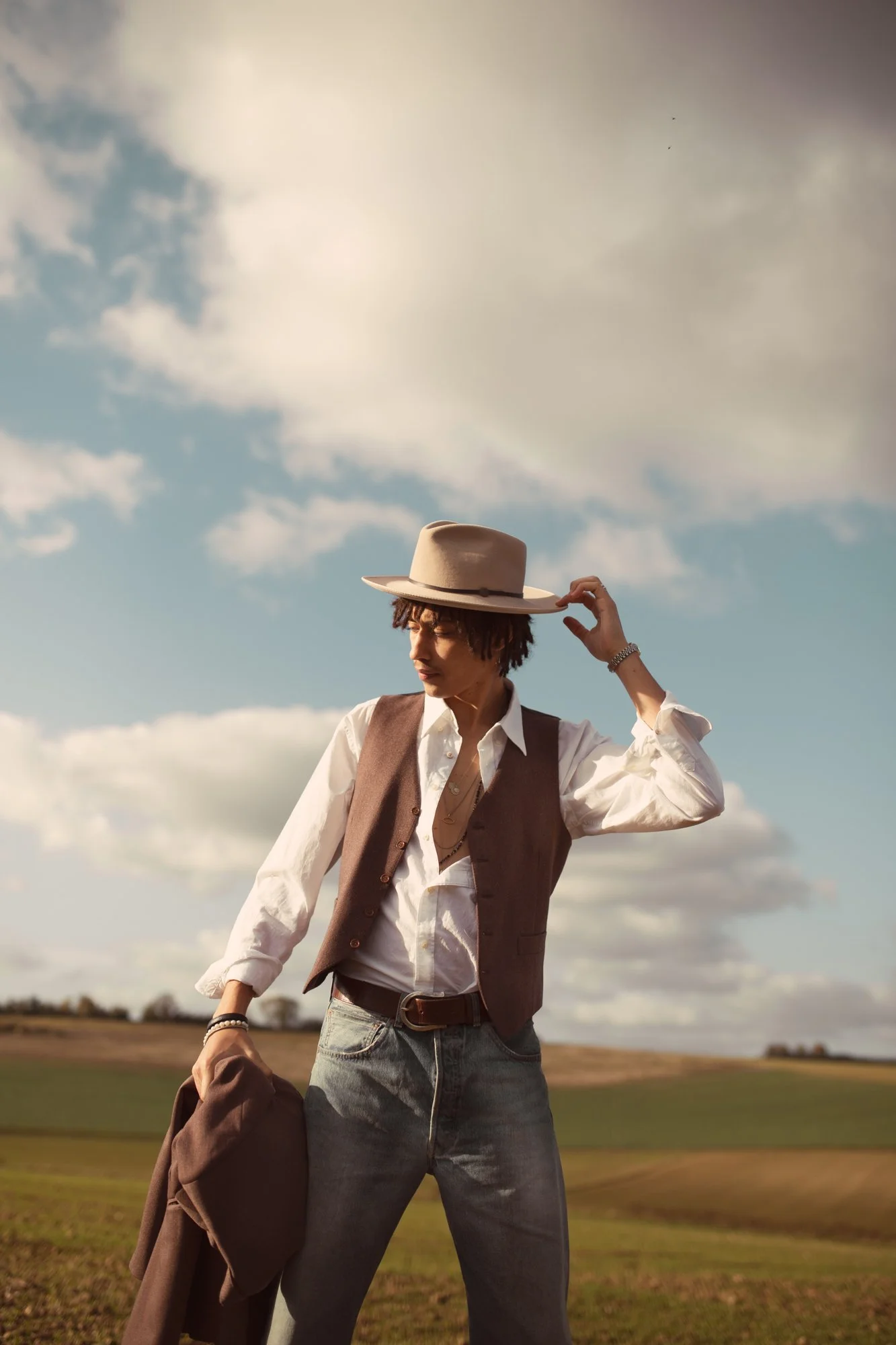 Jeune homme dans une tenue décontractée tenant un manteau marron et portant un chapeau beige, en plein air sous un ciel nuageux.
