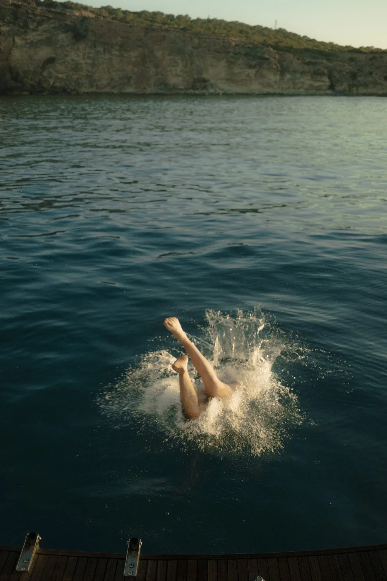 Une personne est en train de plonger dans l'eau depuis une plateforme en bois, créant un splash. La scène se déroule au bord de l'eau avec des rochers en arrière-plan à majorque