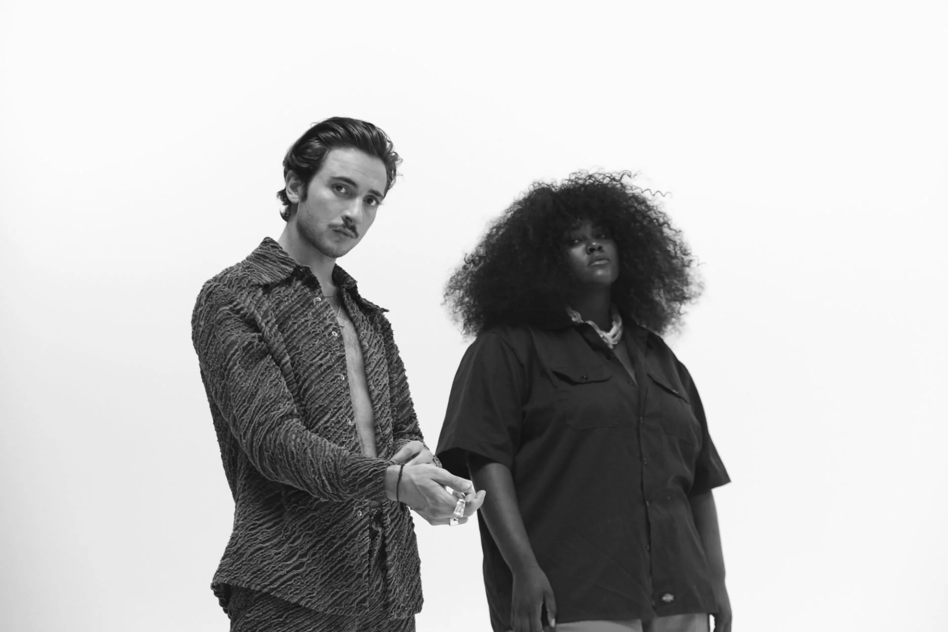 Lord Esperanza & Yseult. L'homme à gauche a les cheveux noirs, porte une chemise à motifs et regarde la caméra. La femme à droite a de grands cheveux bouclés, porte une veste noir et regarde hors du cadre.