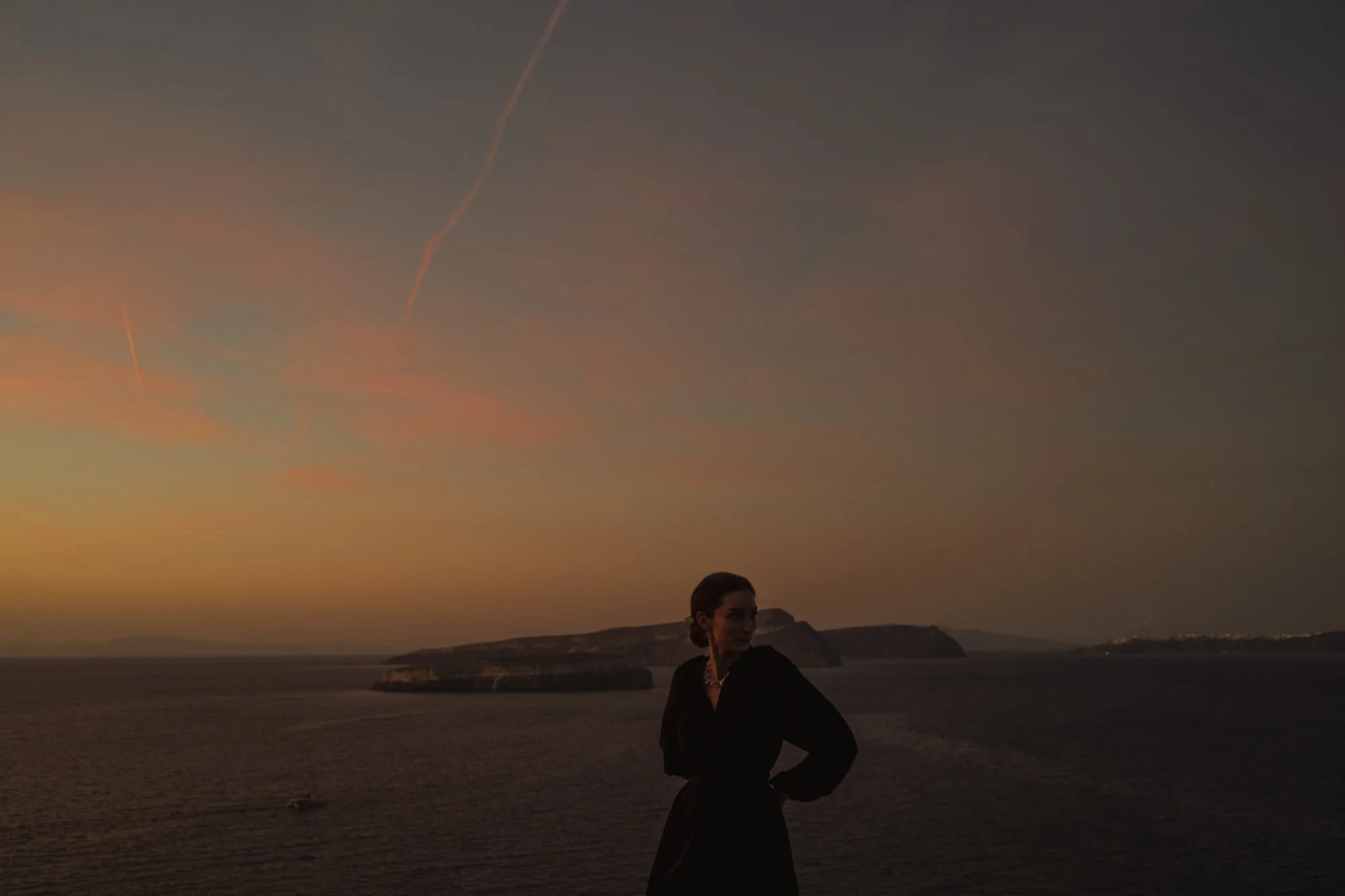 Une femme se tient sur une plage au coucher du soleil, avec un ciel teinté de couleurs chaudes et des formations rocheuses en arrière-plan à santorin