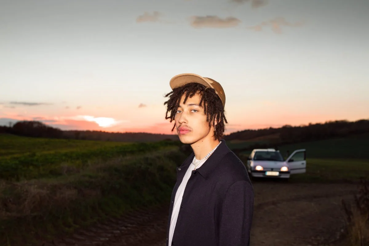 Un jeune homme avec des dreadlocks porte une casquette beige et un manteau noir dans un paysage rural au coucher du soleil, avec une voiture blanche en arrière-plan.
