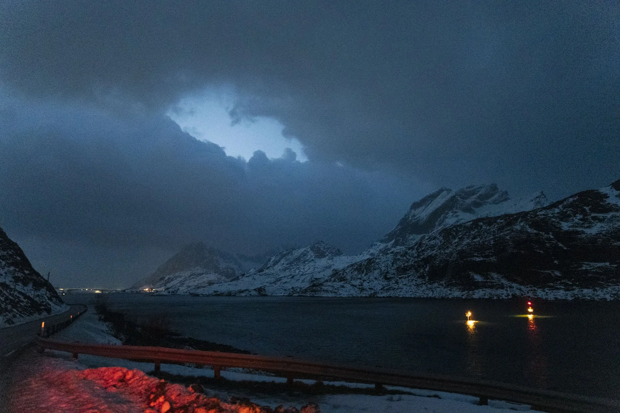 Montagnes enneigées sous un ciel nuageux avec une ouverture illuminée, reflet de lumières sur l'eau, route sinueuse avec barrières en bois, ambiance nocturne.»