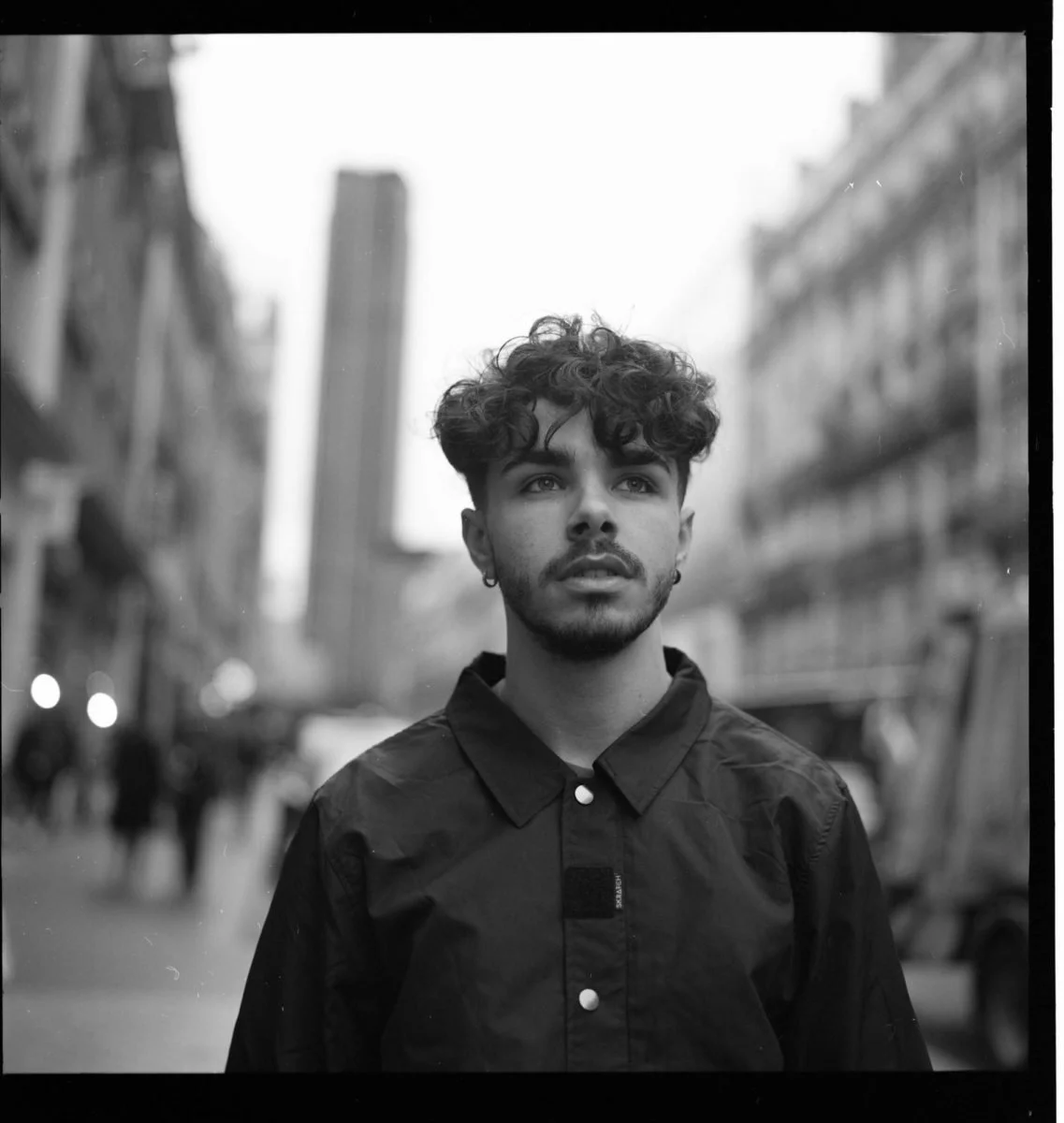 Un jeune homme avec des cheveux bouclés et une barbe, portant une chemise noire, se tient dans une rue urbaine avec des bâtiments en arrière-plan. L'image est en noir et blanc.