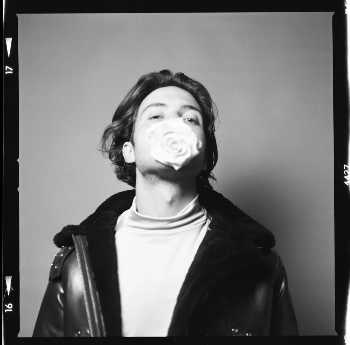Un homme avec des cheveux longs et ondulés, portant un manteau en cuir et un t-shirt blanc, avec un visage partiellement obscurci par une rose, photo en noir et blanc.