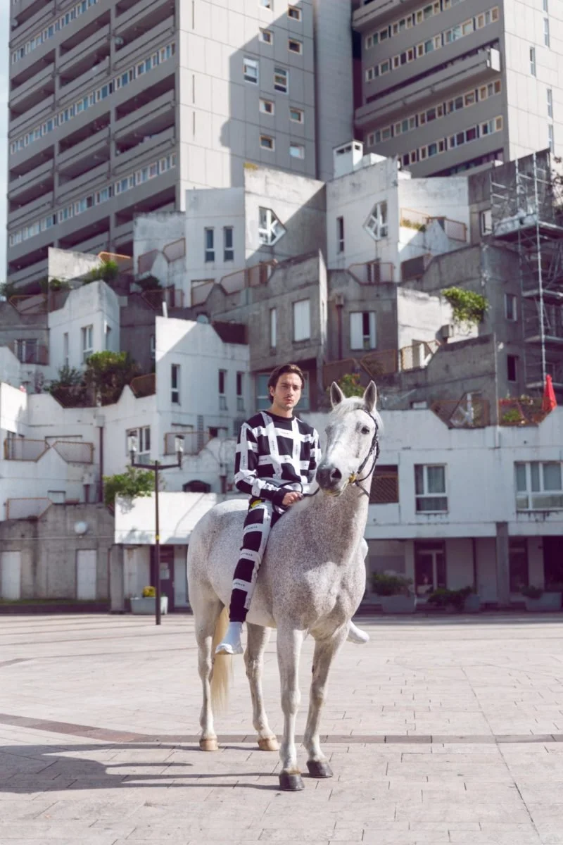 Un homme en vêtements noirs et blancs assis sur un cheval blanc dans une place urbaine, avec un bâtiment moderne en arrière-plan.