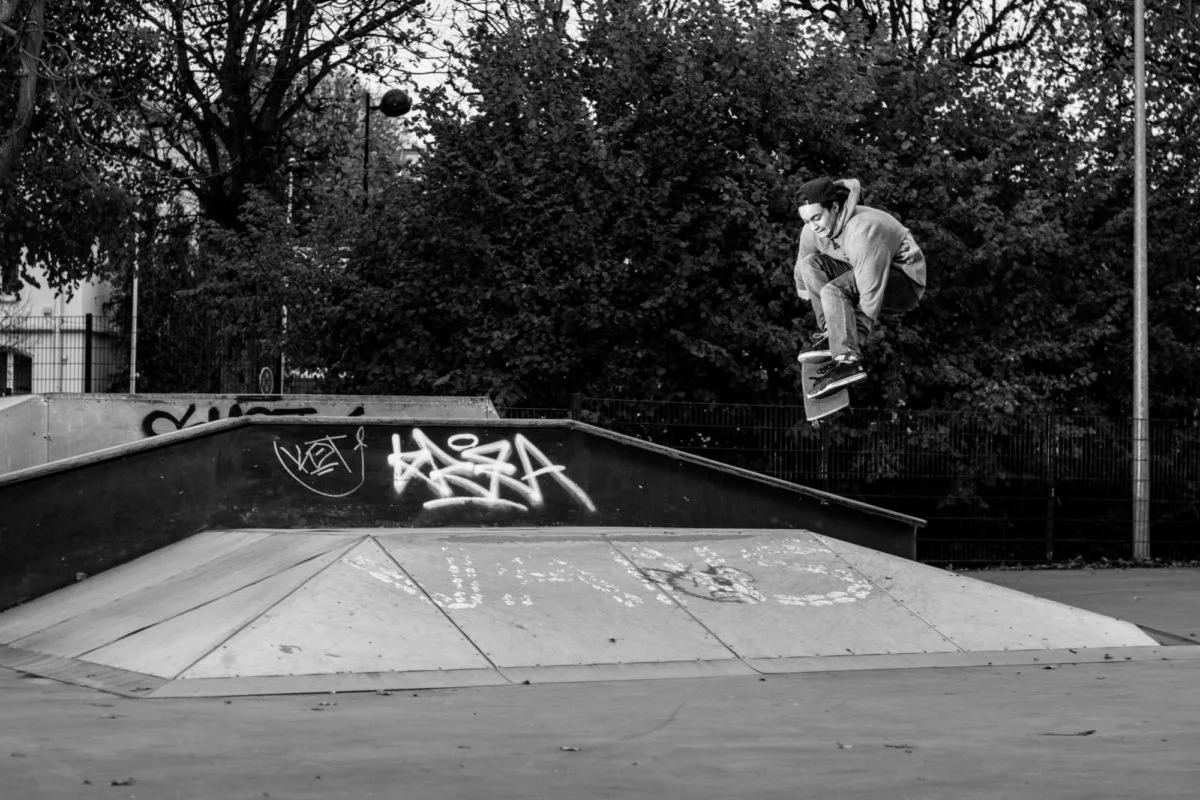 Un skateur en plein saut dans un skatepark, avec des graffitis sur la rampe, entouré d'arbres et de lampadaires en arrière-plan, en noir et blanc.