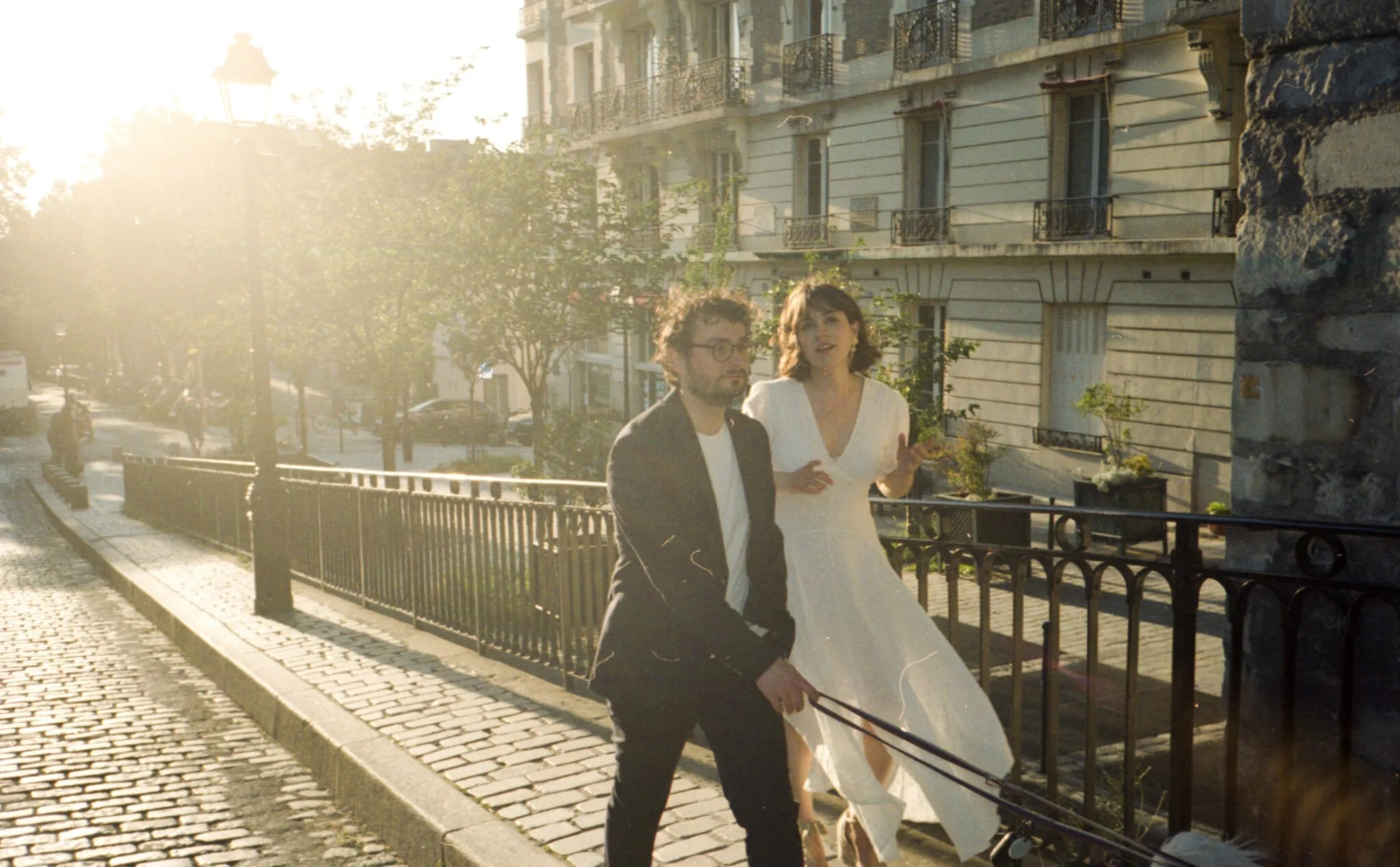 Un couple, un homme avec des lunettes et une femme en robe blanche, marche sur un trottoir en ville sous un soleil couchant, avec des bâtiments anciens en arrière-plan.
