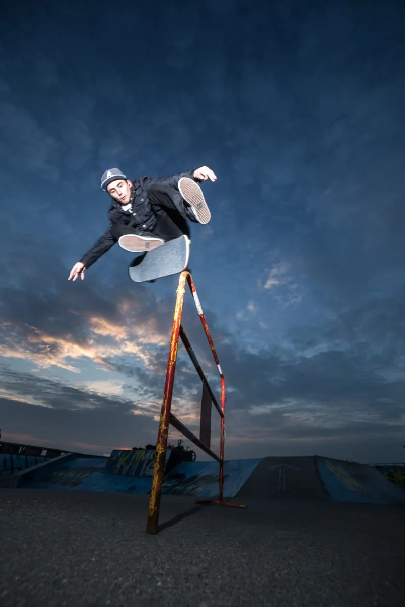 Joseph Garbaccio en train de faire un trick aérien sur une rampe dans un skatepark au coucher du soleil, ciel nuageux.