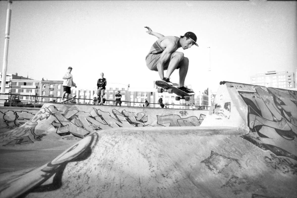 Un skateur en train de sauter dans un skatepark urbain, avec d'autres personnes en arrière-plan et des bâtiments modernes.