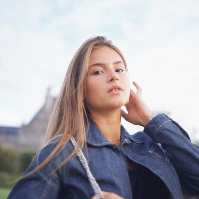 Jeune femme avec des cheveux longs et blonds, portant une veste en denim, posé en extérieur avec un bâtiment flou en arrière-plan.