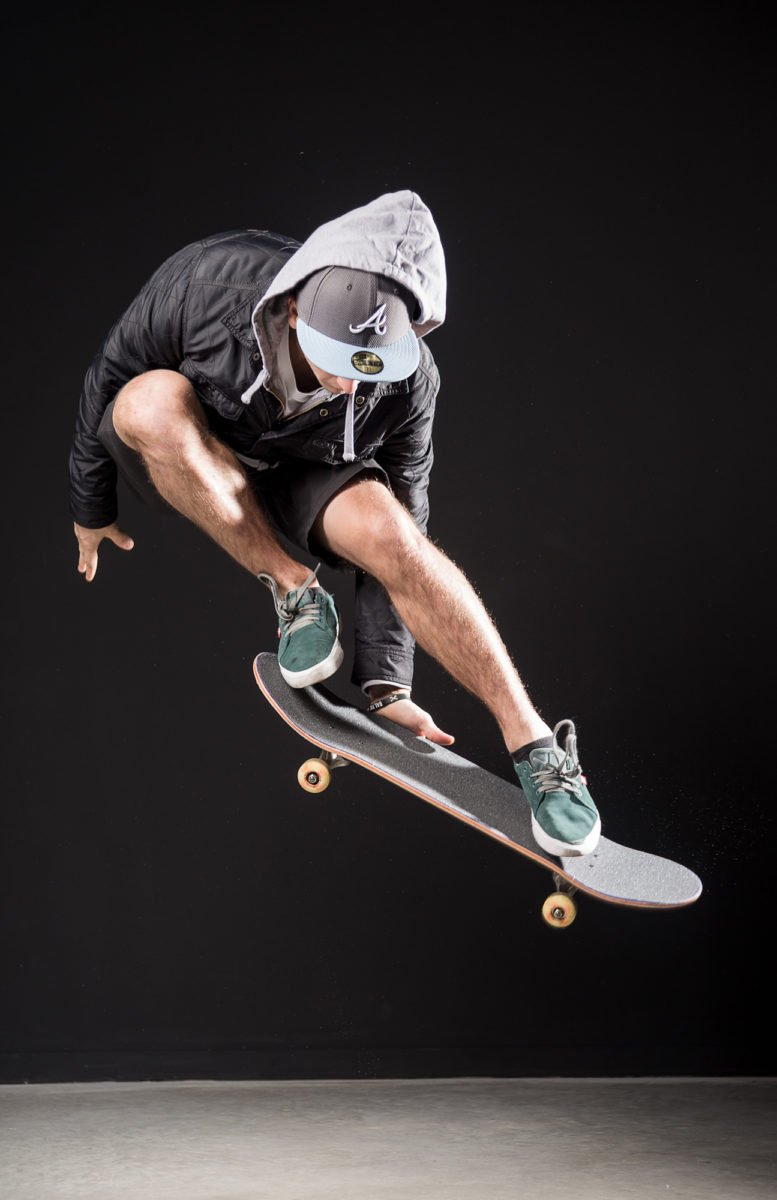 Joseph Garbaccio en hoodie, casquette et shorts pratique le skateboard en sautant dans un espace intérieur avec un fond noir.