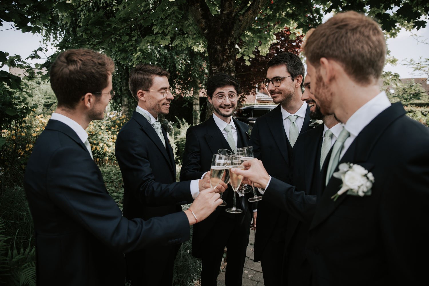 Groupe de six hommes en costume noir, célébrant avec des flûtes de champagne lors d'une occasion spéciale en plein air, sous un arbre.