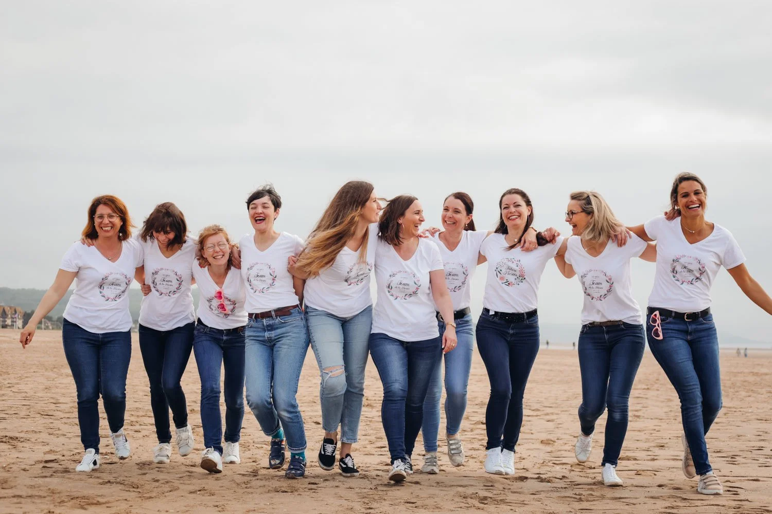 Groupe de neuf femmes souriantes et riant en marchant sur la plage, portant des t-shirts blancs avec un motif floral et le texte 'team', et des jeans, sous un ciel nuageux.
