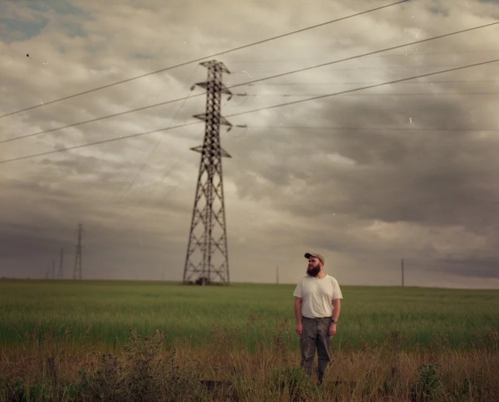 Un homme avec une barbe, portant un t-shirt blanc et un chapeau, se tient dans un champ vert sous un ciel nuageux avec des lignes électriques en arrière-plan.