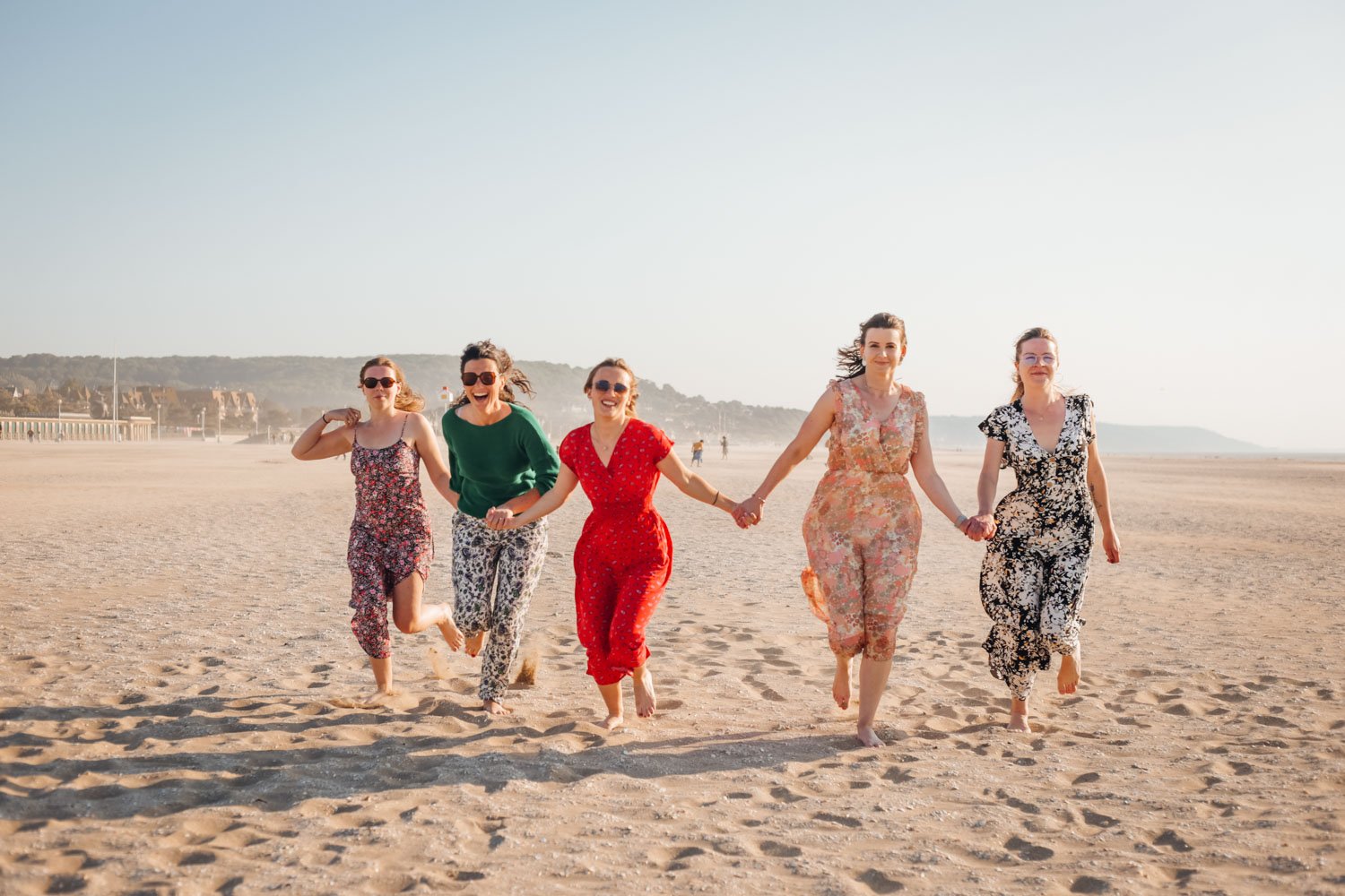 Cinq femmes en robe de couleurs variées, tenant la main, courent sur la plage en souriant avec un ciel clair