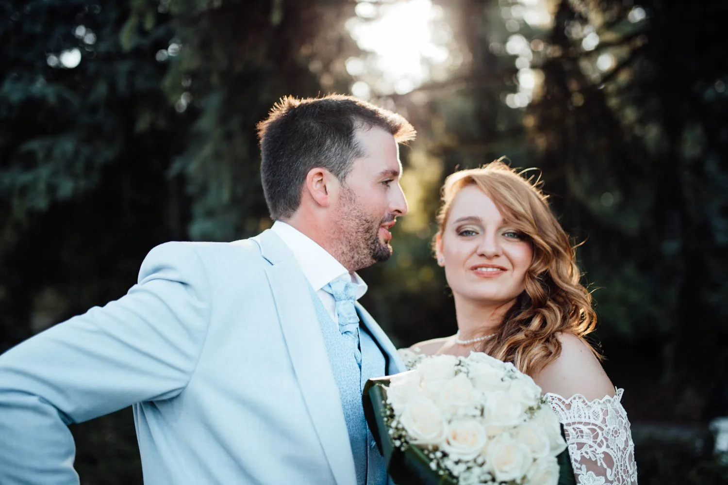 Un couple de mariés lors d'une séance photo en plein air, avec des arbres en arrière-plan, la femme tient un bouquet de roses blanches