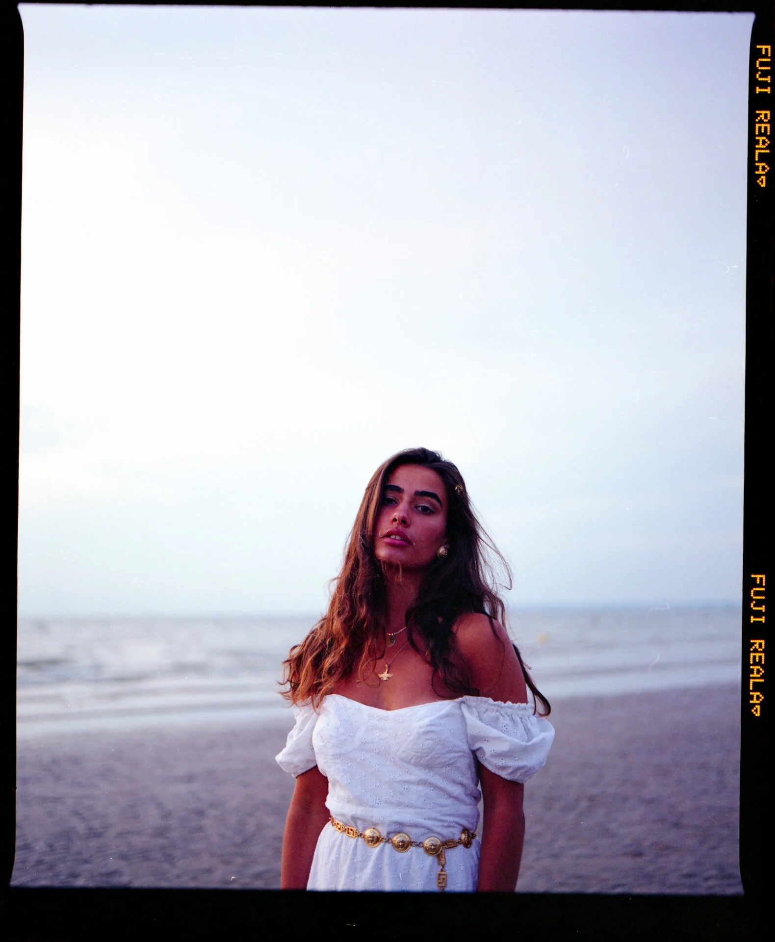 Une jeune femme avec de longs cheveux bruns portant une robe blanche, avec des bijoux dorés, se tient sur la plage au coucher du soleil.