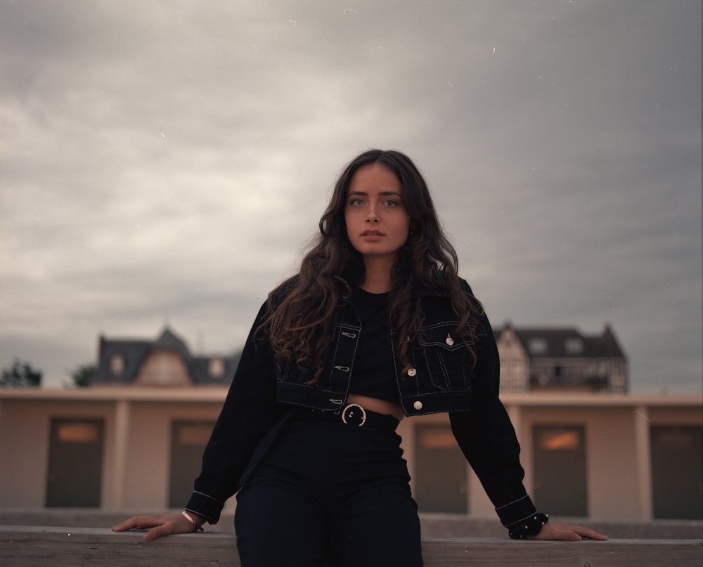 Jeune femme aux cheveux longs et bouclés, portant un crop top noir et une veste en denim noire, posant sur un pont avec un ciel nuageux en arrière-plan.