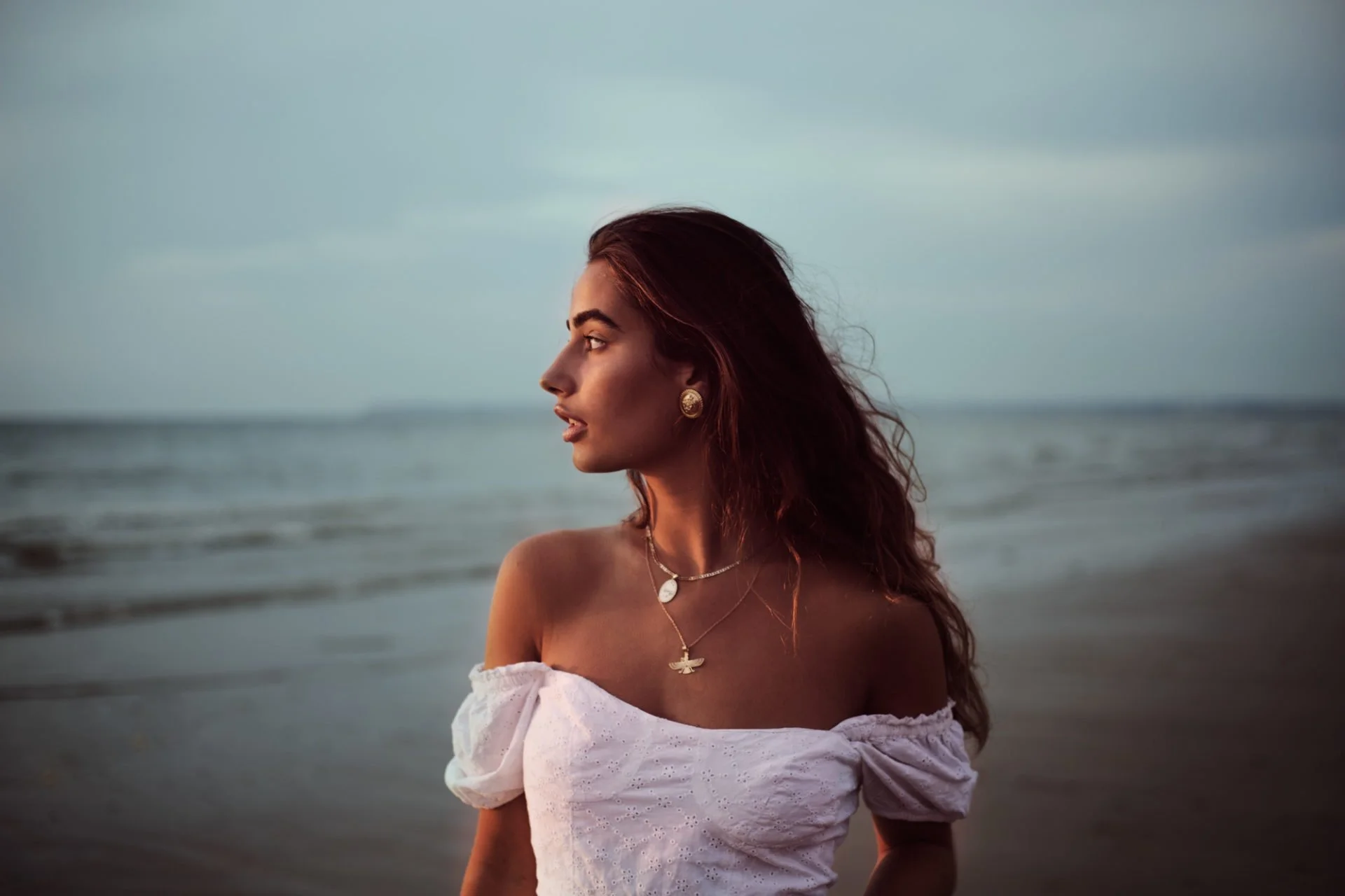 Femme aux cheveux bruns portant des bijoux, regardant vers la gauche, sur une plage au coucher du soleil.