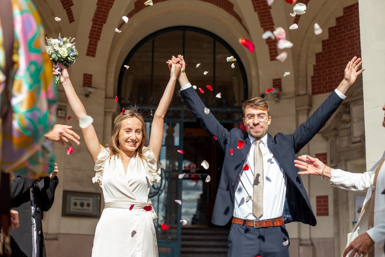 Un couple de mariés souriants après leur mariage, levant les bras en l'air sous des pétales de fleurs tombant du ciel, en dehors d'une église