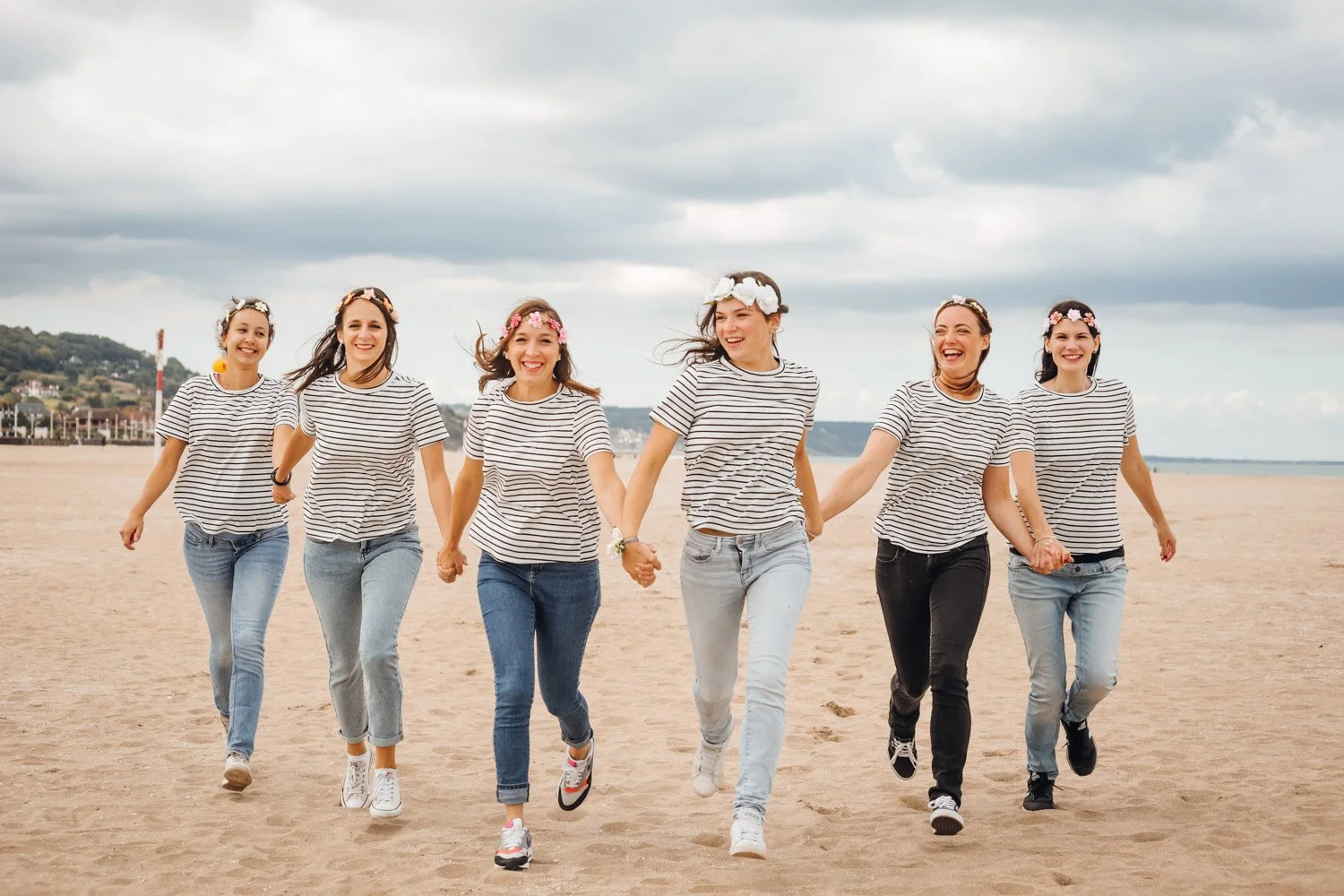 Groupe de six jeunes femmes marchant et souriant sur une plage, habillées en rayures et portant des couronnes de fleurs.