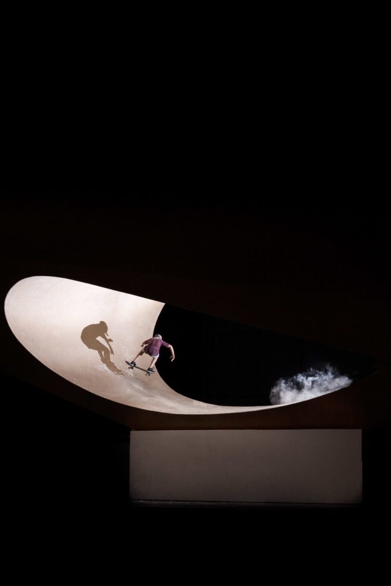 Un skateur skate dans un bowl en béton à l'intérieur d'une structure avec un éclairage dramatique.