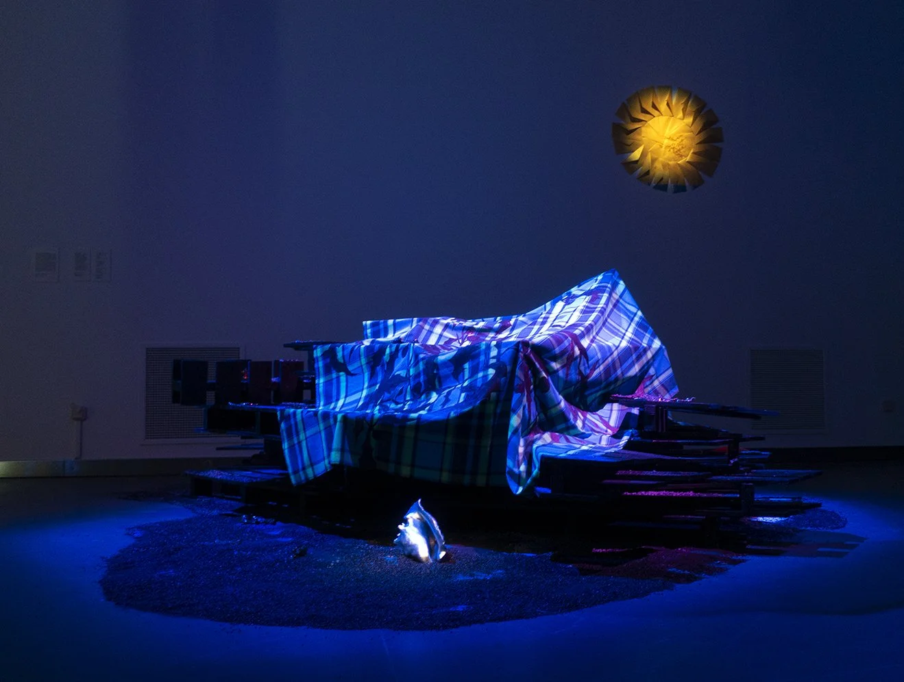 Installation artistique avec un ensemble de palettes en bois sur lesquelles un tissu madras est posé. L'installation dispose d'un éclairage bleu et d'un soleil sur le mur, réalisé en enveloppes dorées.