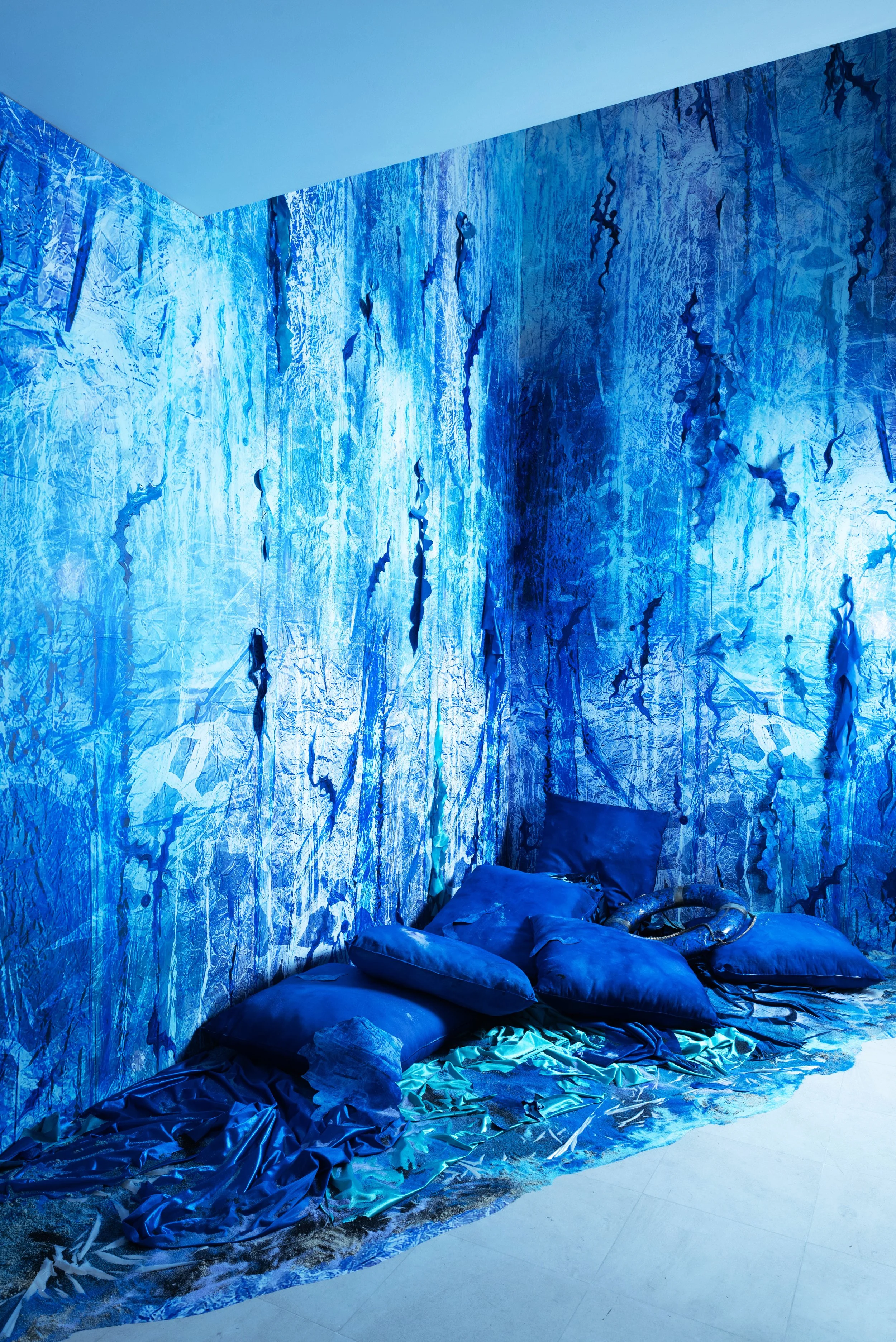 Une pièce avec des murs recouverts de photographies aux nuances de bleu, avec des coussins et des draps également bleu sur le sol, créant une ambiance futuriste ou artistique. C'est une installation spécifique conçue pour la Fondation Blachère.