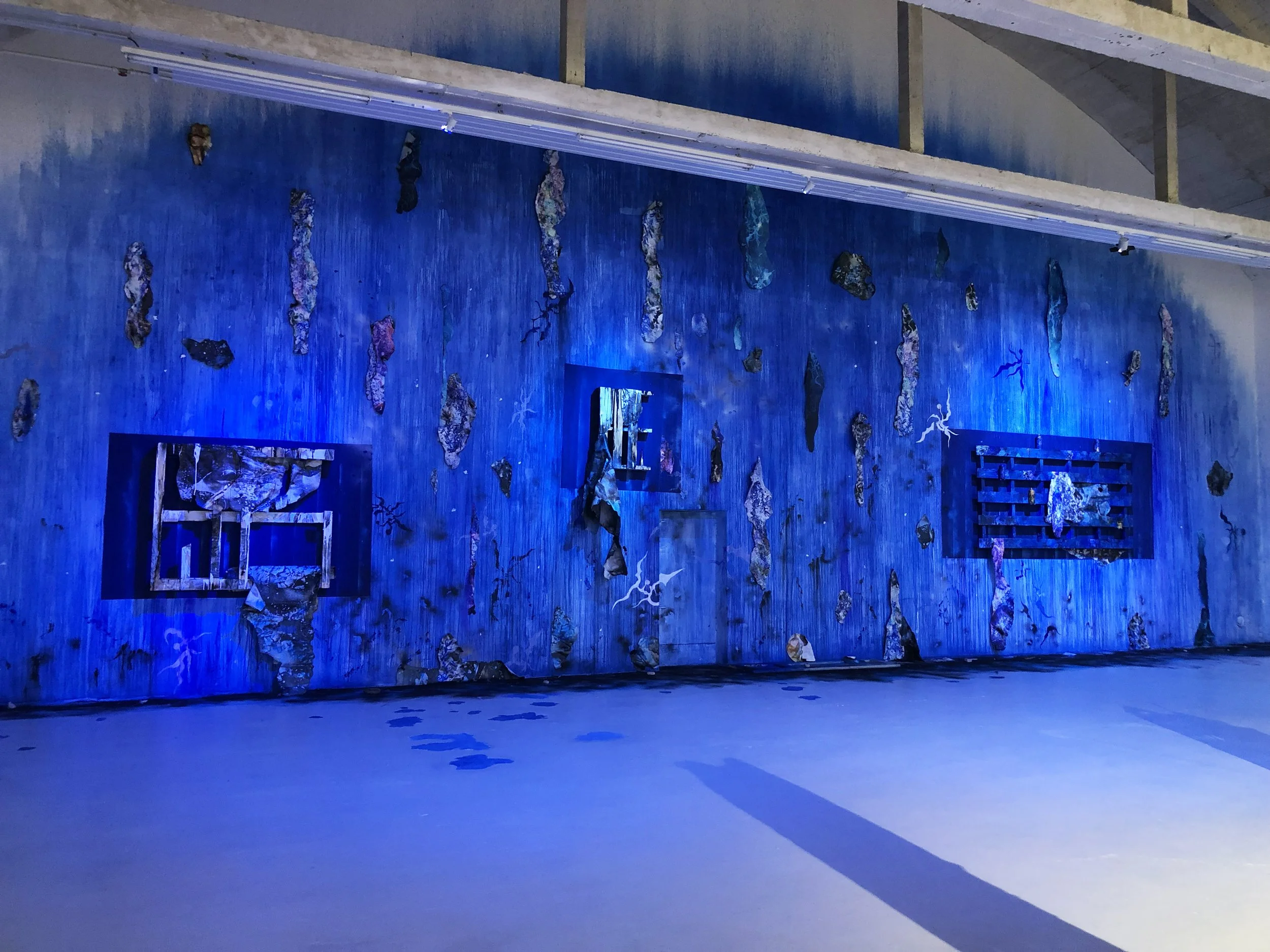 Une installation artistique avec un mur peint en bleu avec des morceaux de palettes suspendues, des photographies... Tout cela dans une ambiance bleue.