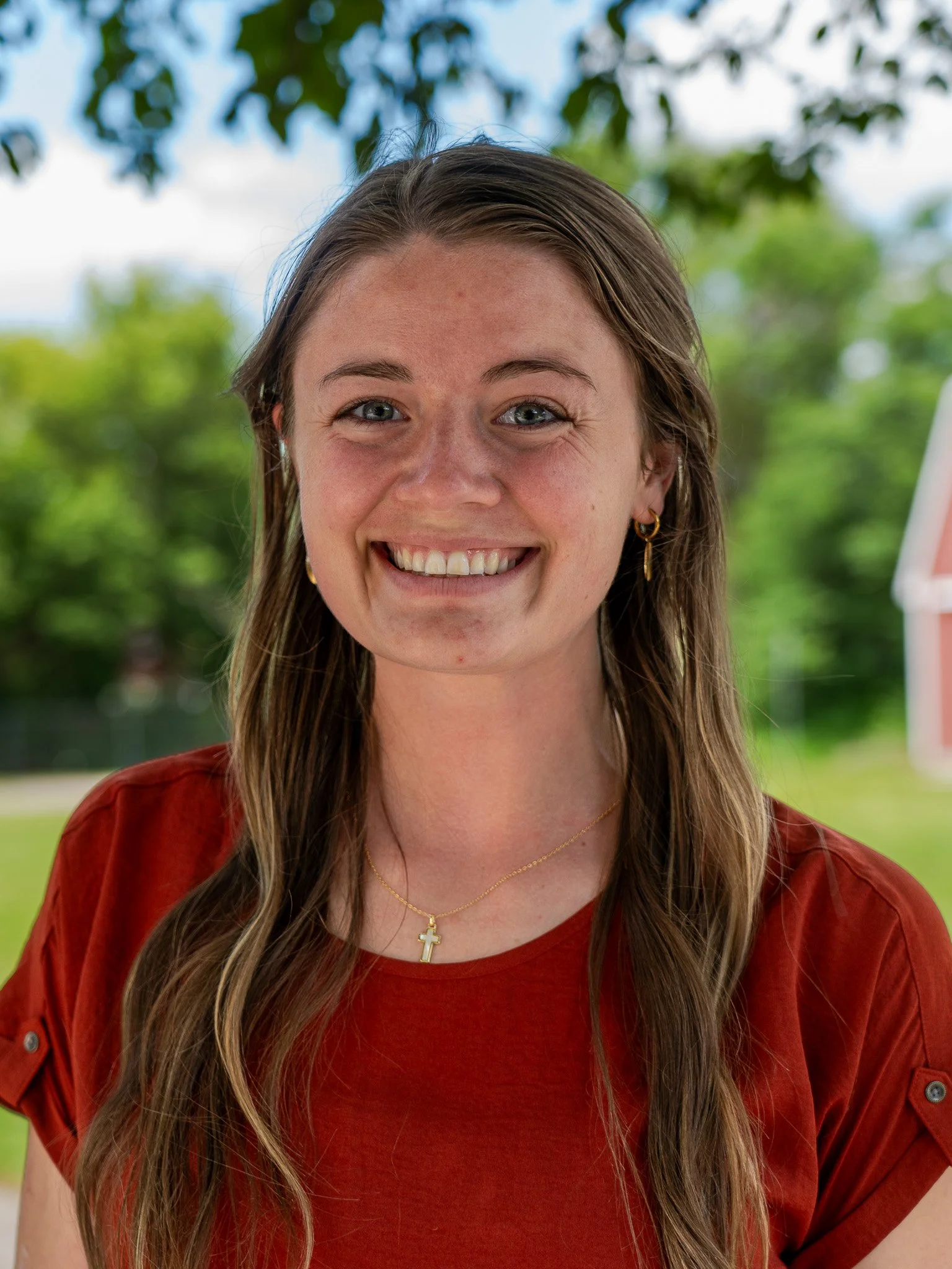 A young woman with long brown hair, wearing a red shirt, gold earrings, and a gold cross necklace, smiling outdoors with greenery and blue sky in the background.