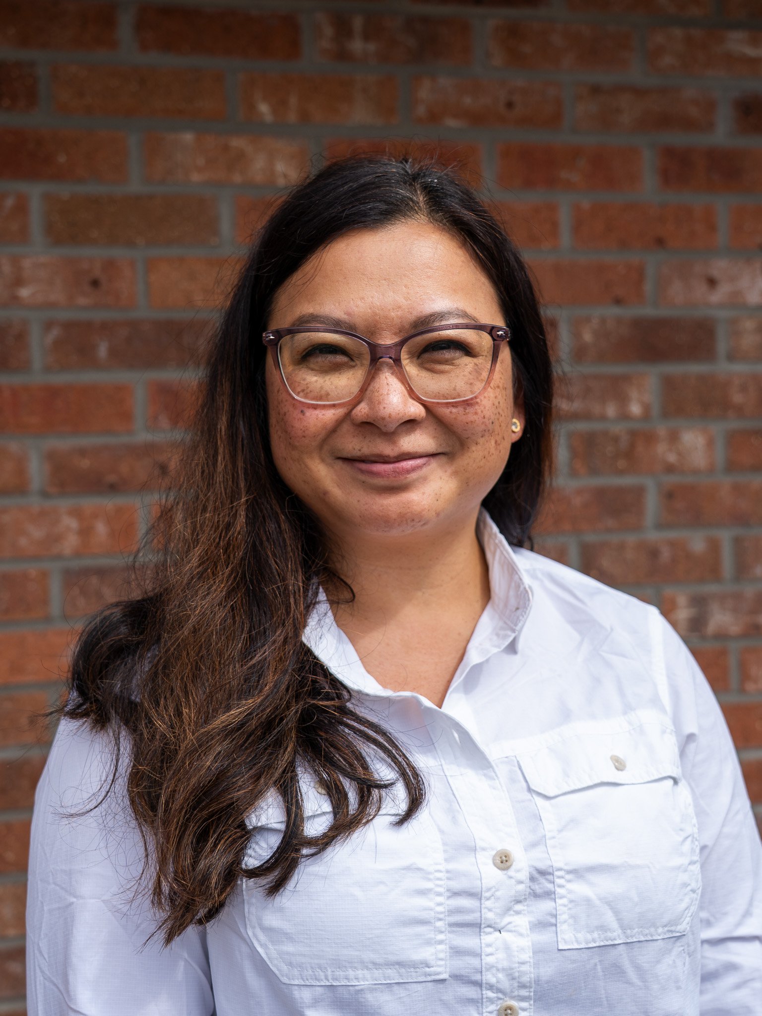 A woman with long dark hair, glasses, and a white button-up shirt standing in front of a brick wall.