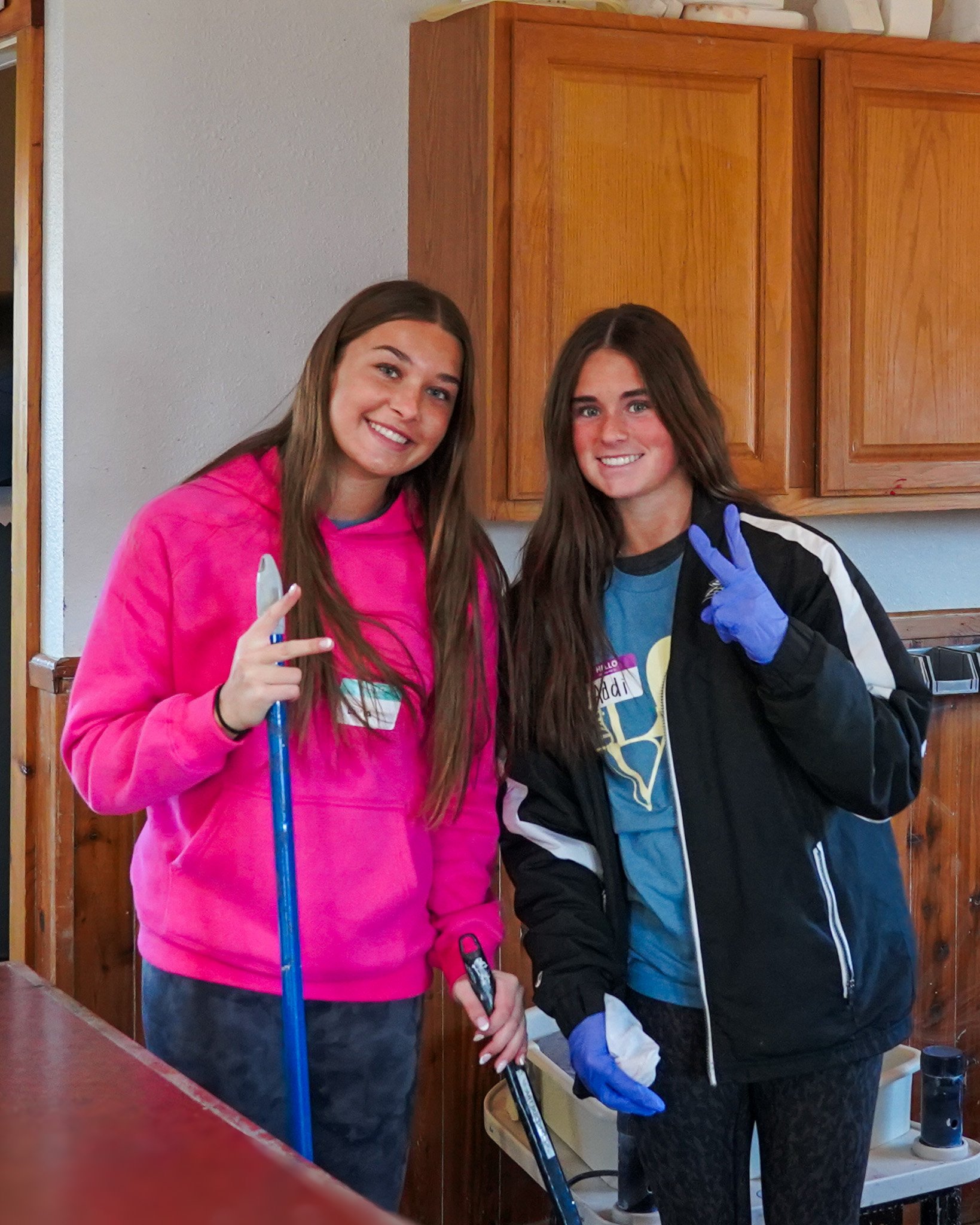 Two young women smiling and posing for a photo in a kitchen. One is wearing a pink hoodie and holding a marker, and the other is wearing a black jacket with blue gloves, making a peace sign.