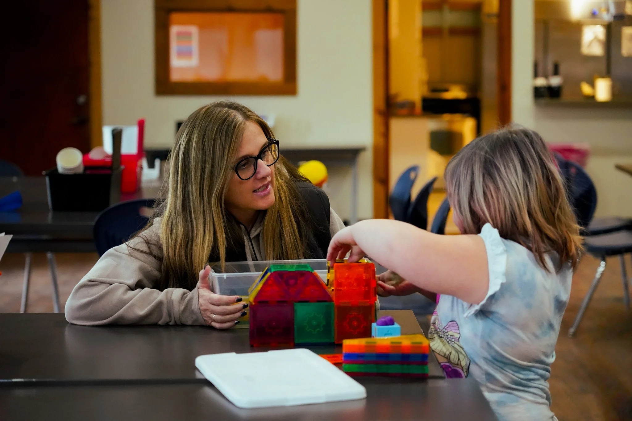 Two women are sitting at a table and playing with colorful magnetic building blocks. One woman is wearing glasses and a beige jacket while the other woman, who has long blonde hair, is wearing a light-colored shirt. The background shows a room with shelves and chairs.