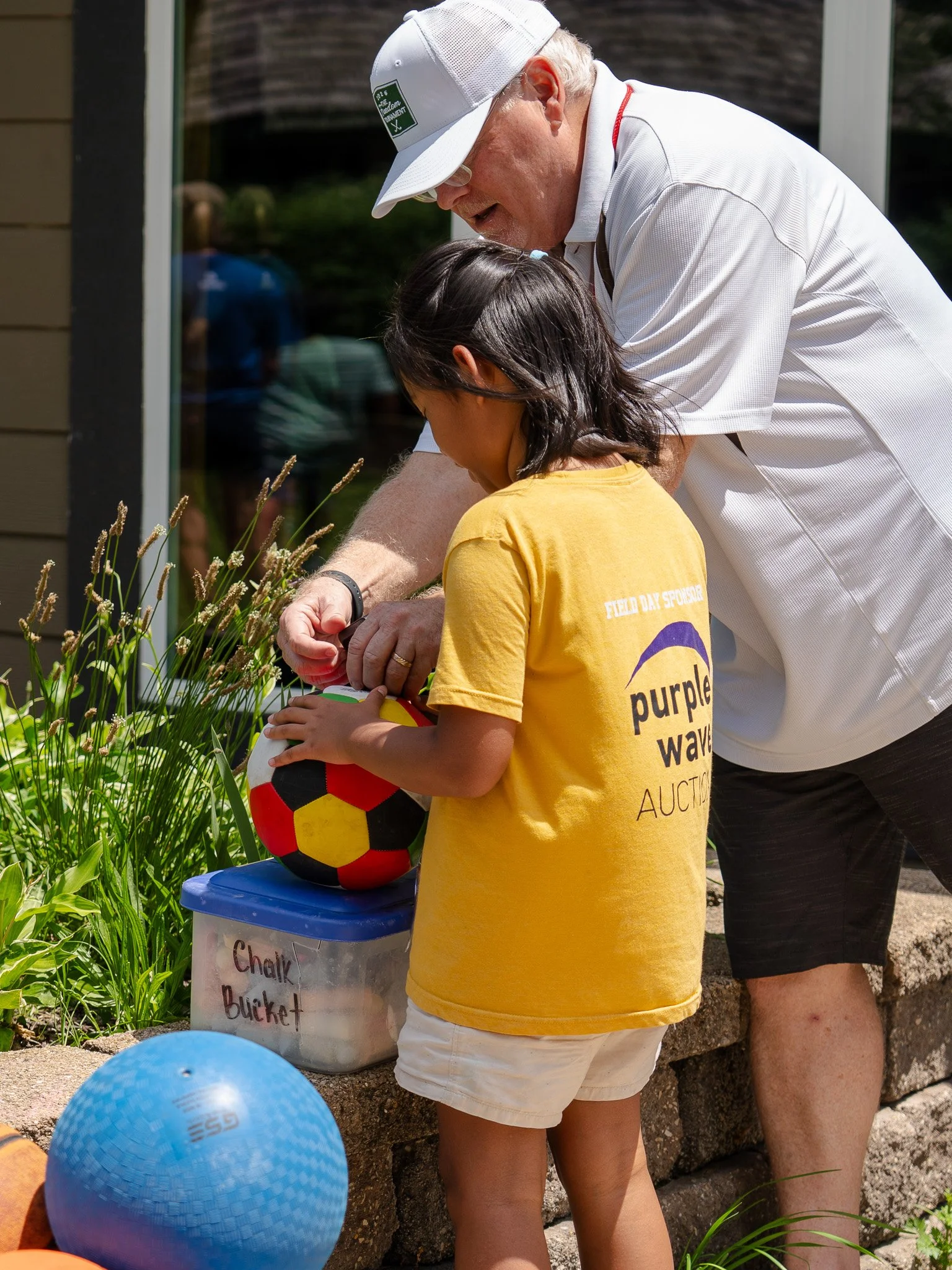 A girl in a yellow T-shirt with 'purple wave auction' printed on it holds a colorful volleyball and stands next to a bin labeled 'chalk' and 'bucket'. An older man with white hair, glasses, a white hat, white polo shirt, and black shorts appears to be helping her, standing outdoors on a sunny day near a garden and building.