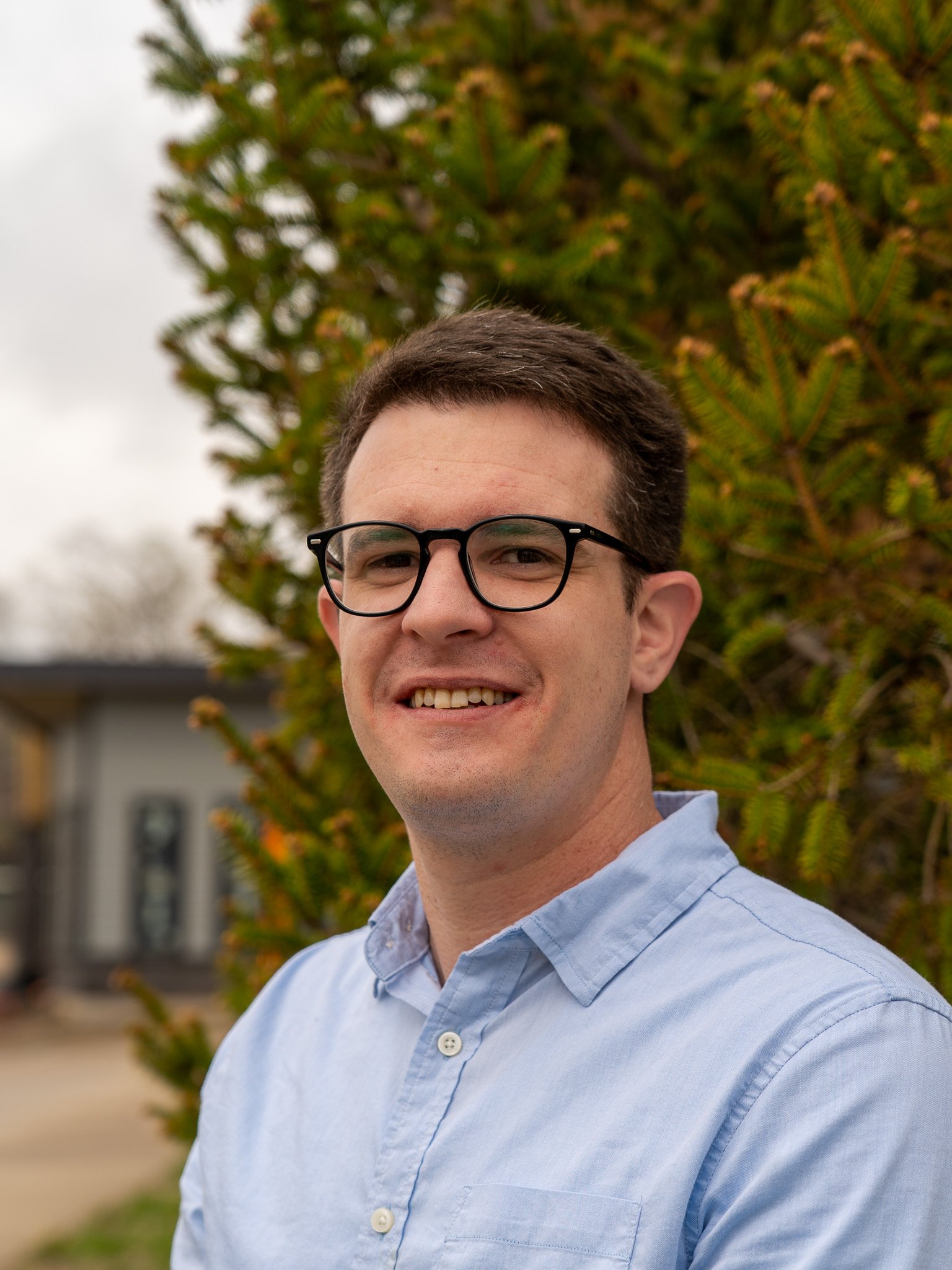 A smiling man with short brown hair, wearing black glasses and a light blue button-up shirt, standing outdoors in front of green shrubbery and a building.