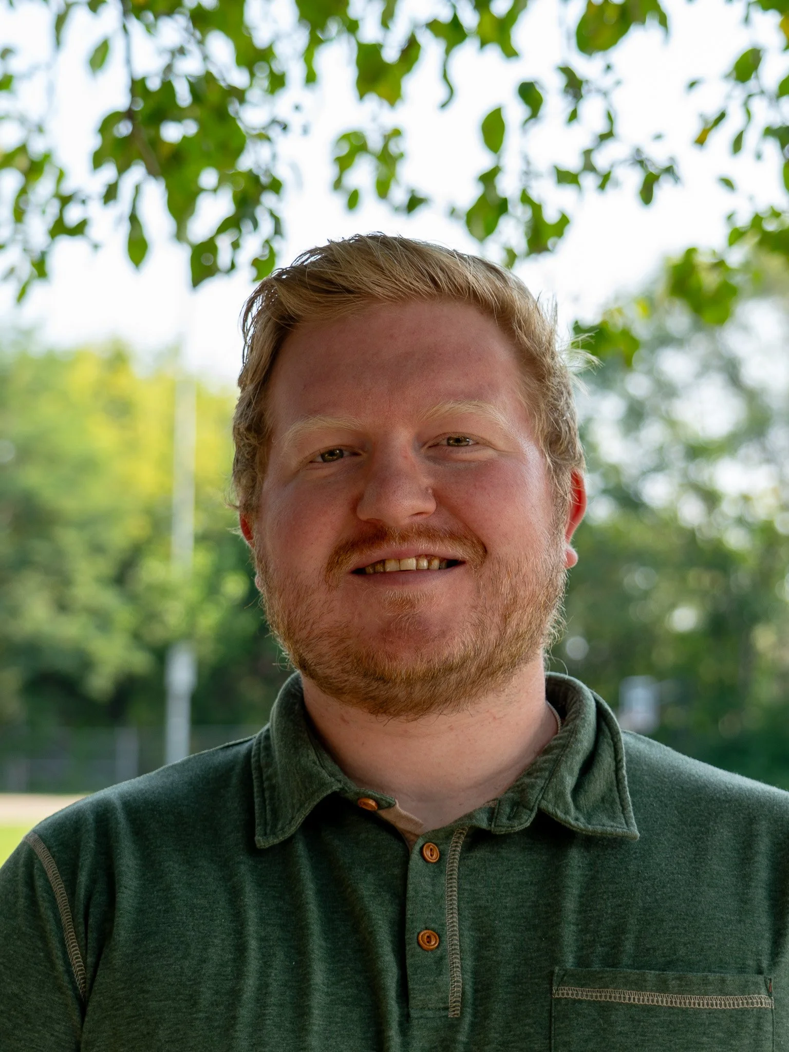 A young man with light skin, reddish hair, and a beard smiling outdoors, wearing a greenish-gray button-up shirt with a collar, with trees and greenery in the background.
