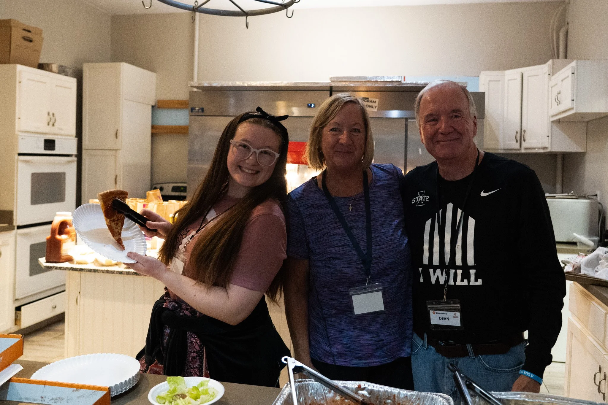 A young woman with glasses and a black headband holding a paper plate with a slice of pizza, standing next to an older woman and a man, in a kitchen. The older woman is wearing a purple shirt, and the man is wearing a black shirt with text. They are smiling, and there are trays of food and paper plates on the counter.