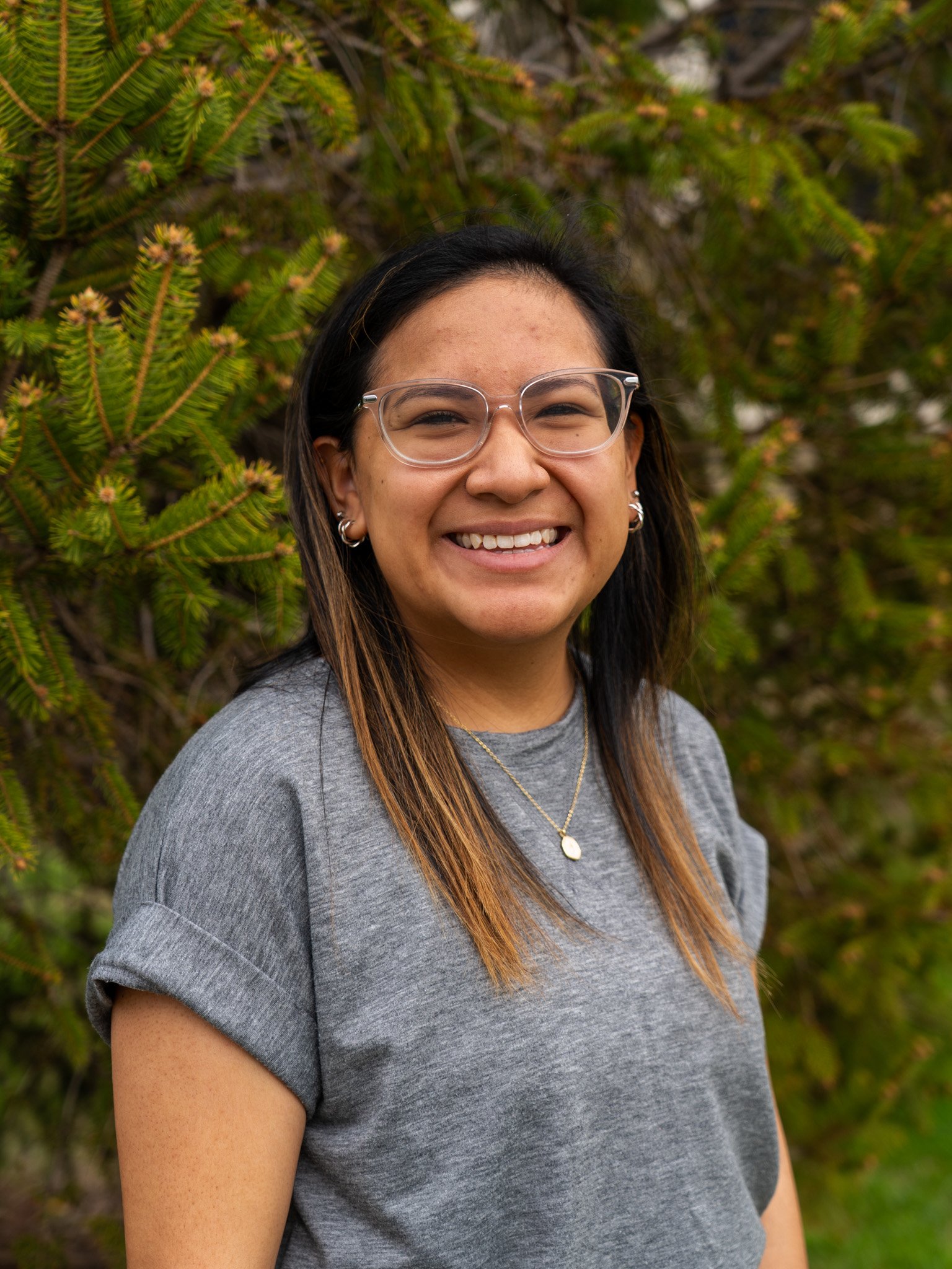 A woman with long hair, wearing glasses and a gray t-shirt, smiling outdoors in front of a green bush.