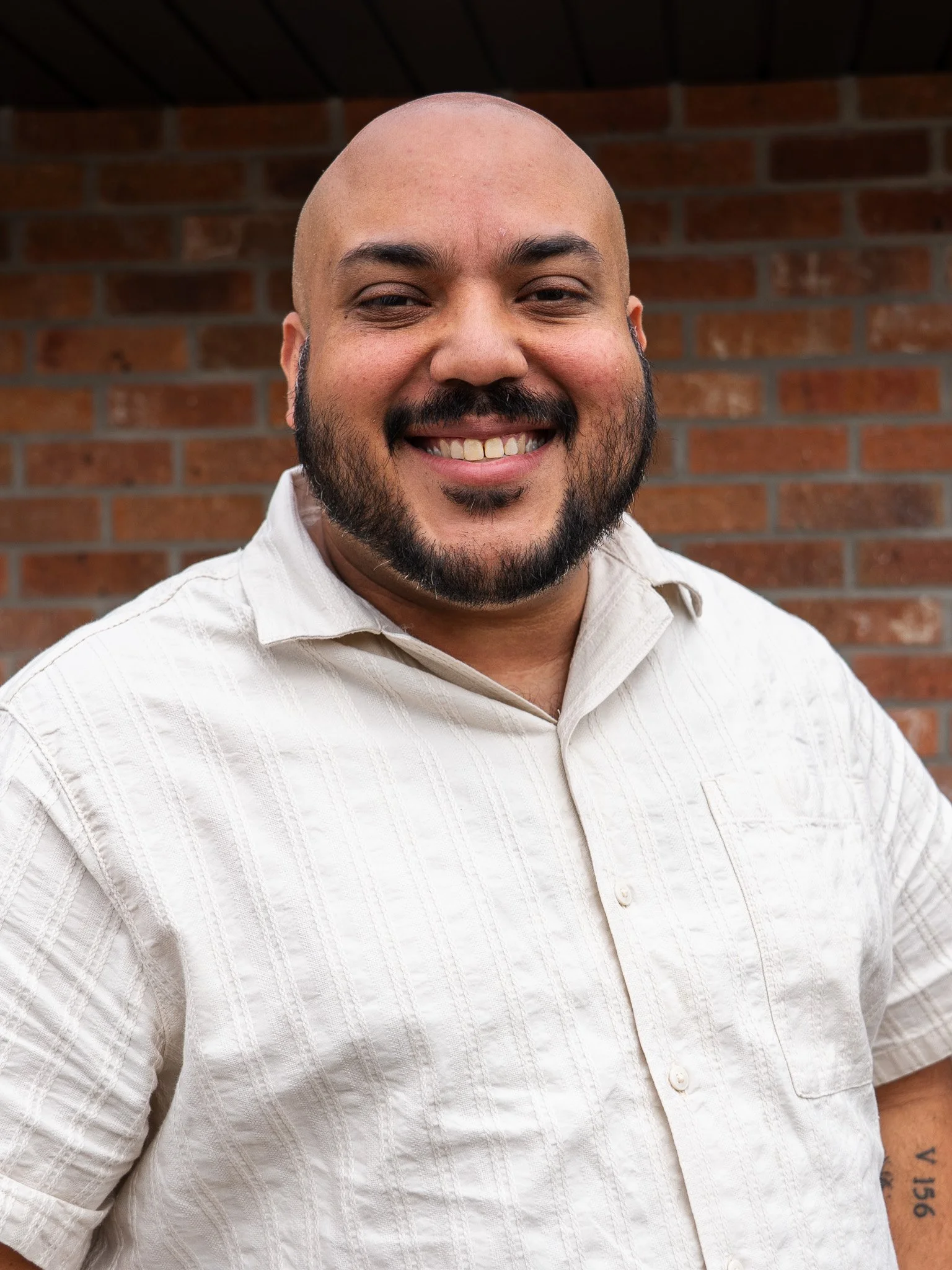 Close-up of a smiling man with a bald head and beard, wearing a cream-colored button-up shirt, standing in front of a brick wall.
