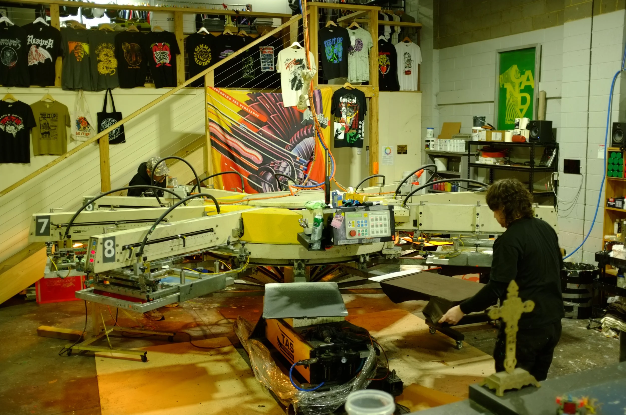 Industrial workshop with machinery, shelves of t-shirts with graphic prints, and a person working on a project. Various tools and equipment are scattered around, with a staircase leading to an upper level in the background.