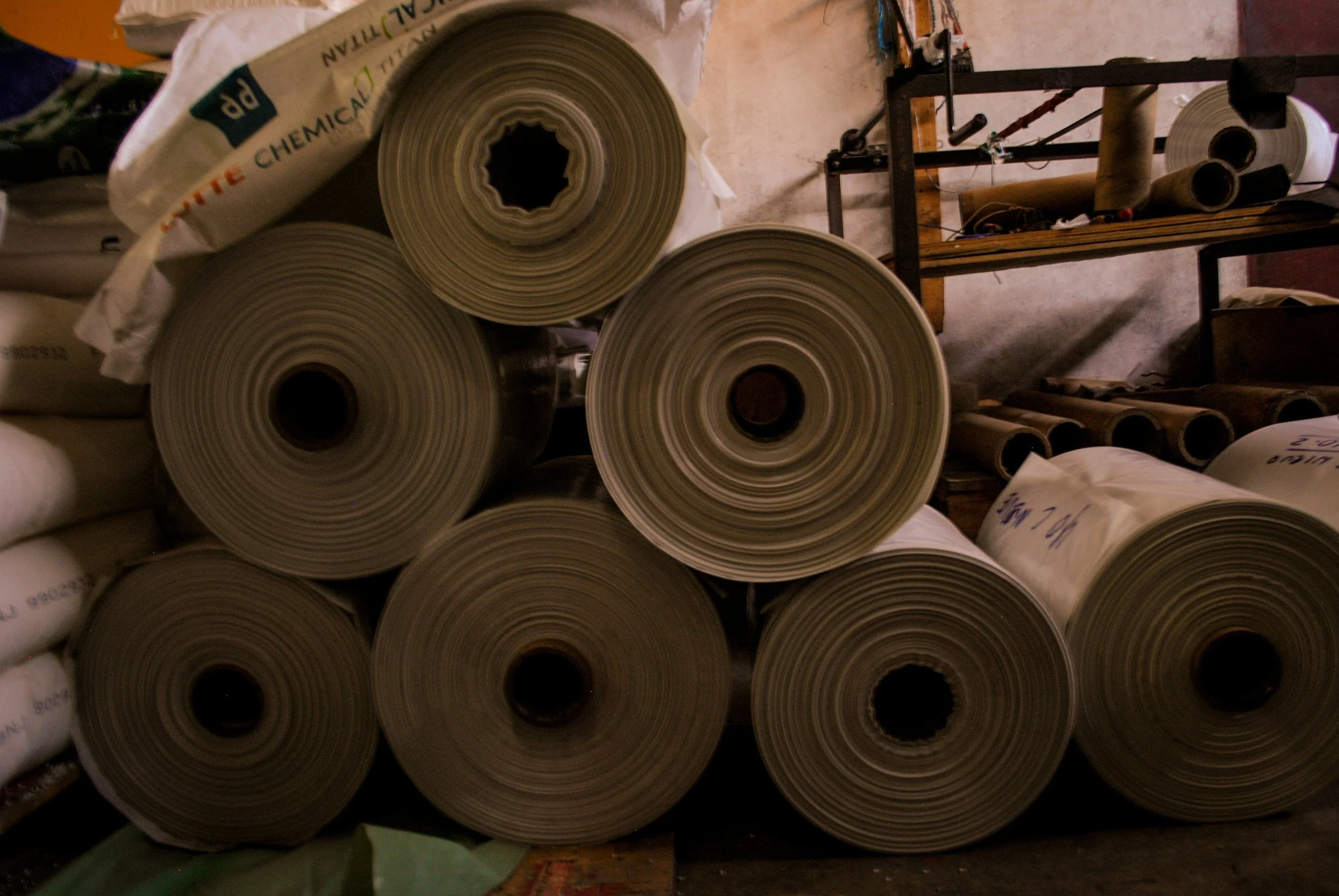 Multiple large rolls of materials, possibly paper or fabric, stacked on each other in a workshop setting with tools and raw materials visible in the background.